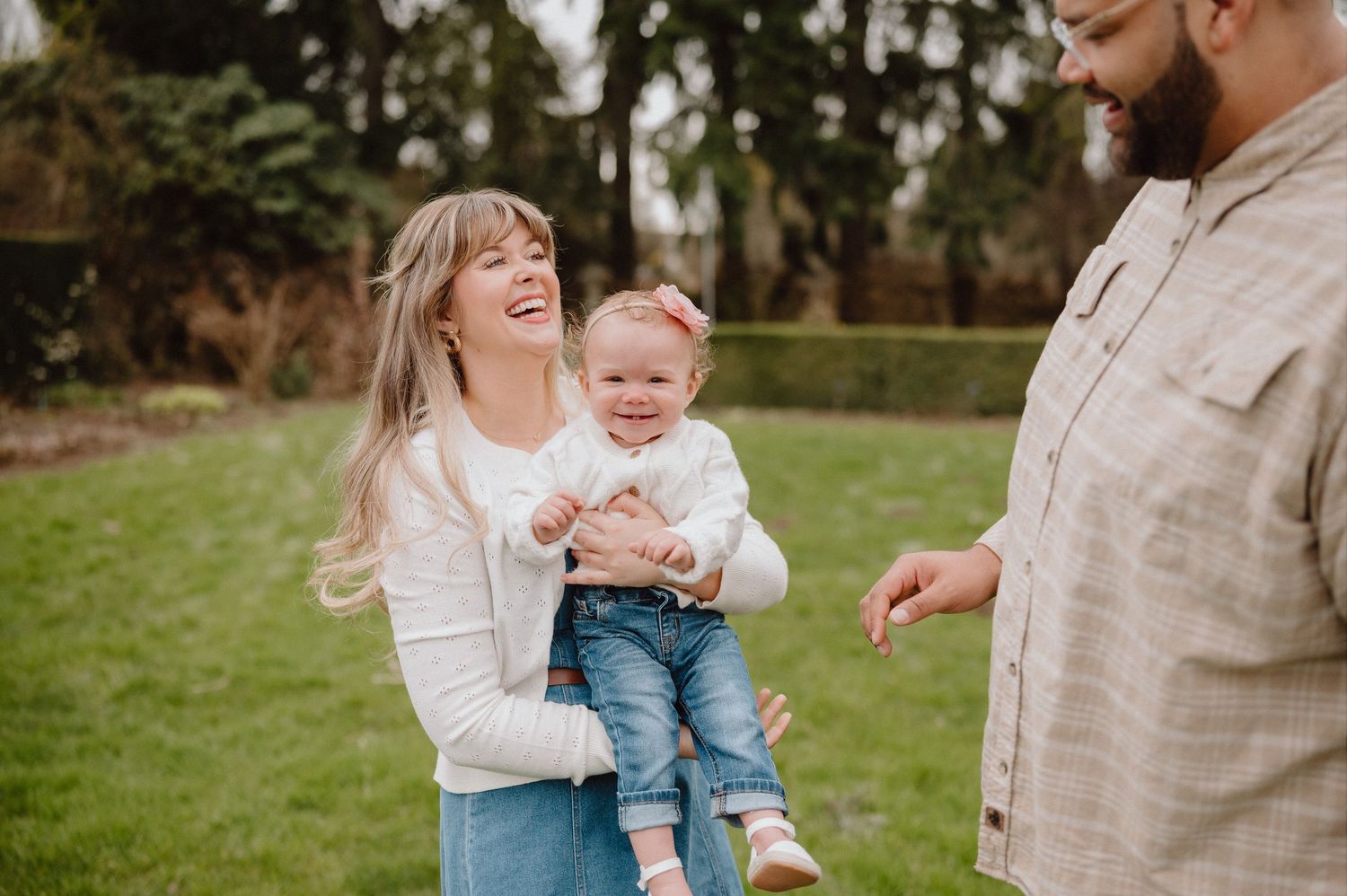 A happy family outdoors, mother holding smiling baby girl while father looks on, in a lush green garden setting.