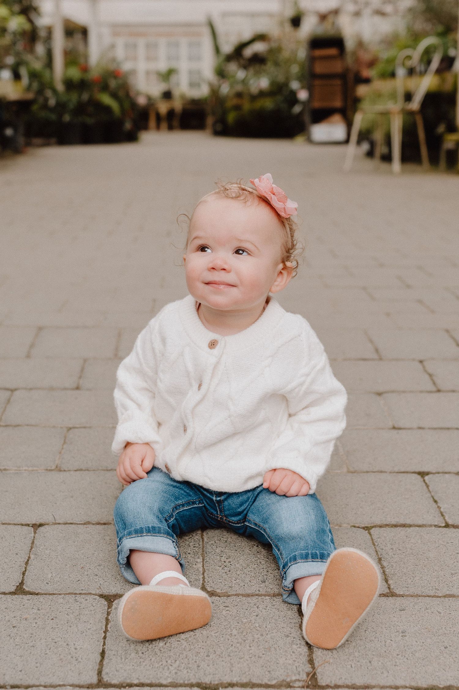 Adorable baby girl in white sweater, jeans, and pink bow sits on stone pavers, smiling upward.