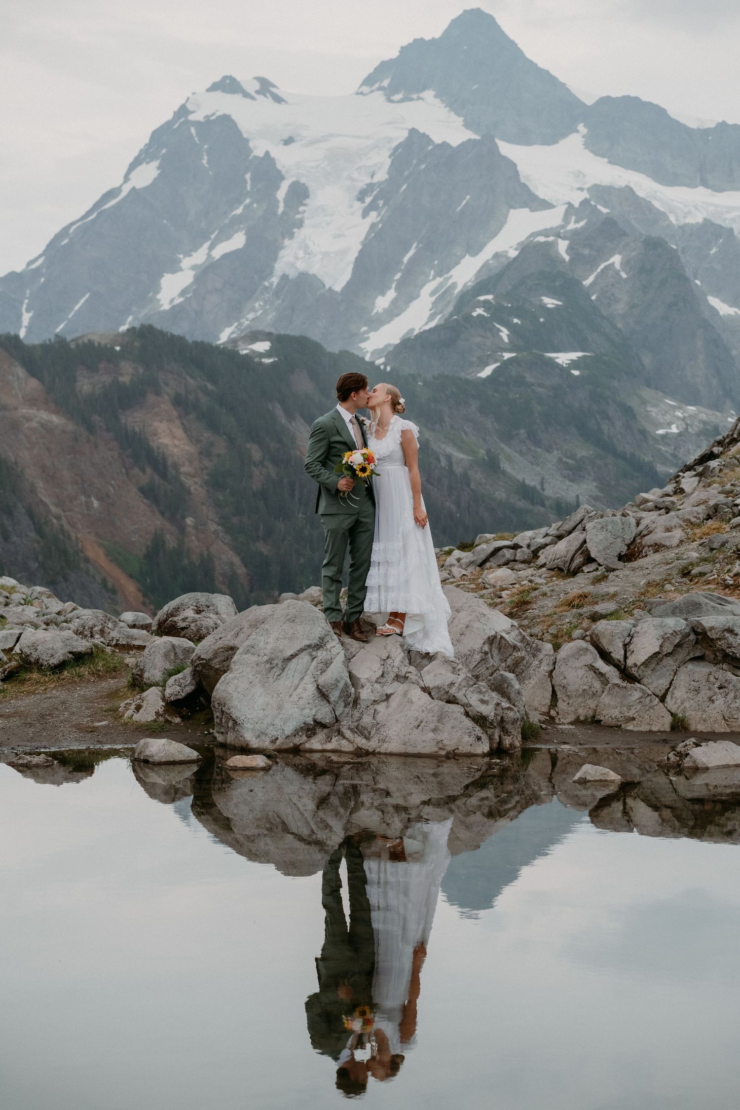A couple embraces near a reflective alpine lake surrounded by snow-capped mountain peaks in a romantic outdoor setting.