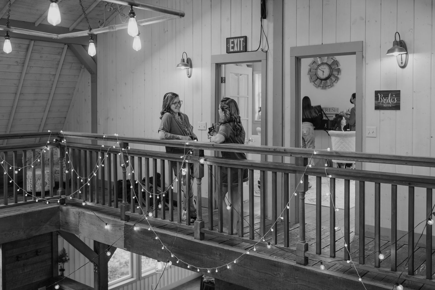 Two women chat on a string-light adorned wooden balcony railing inside a rustic barn venue.