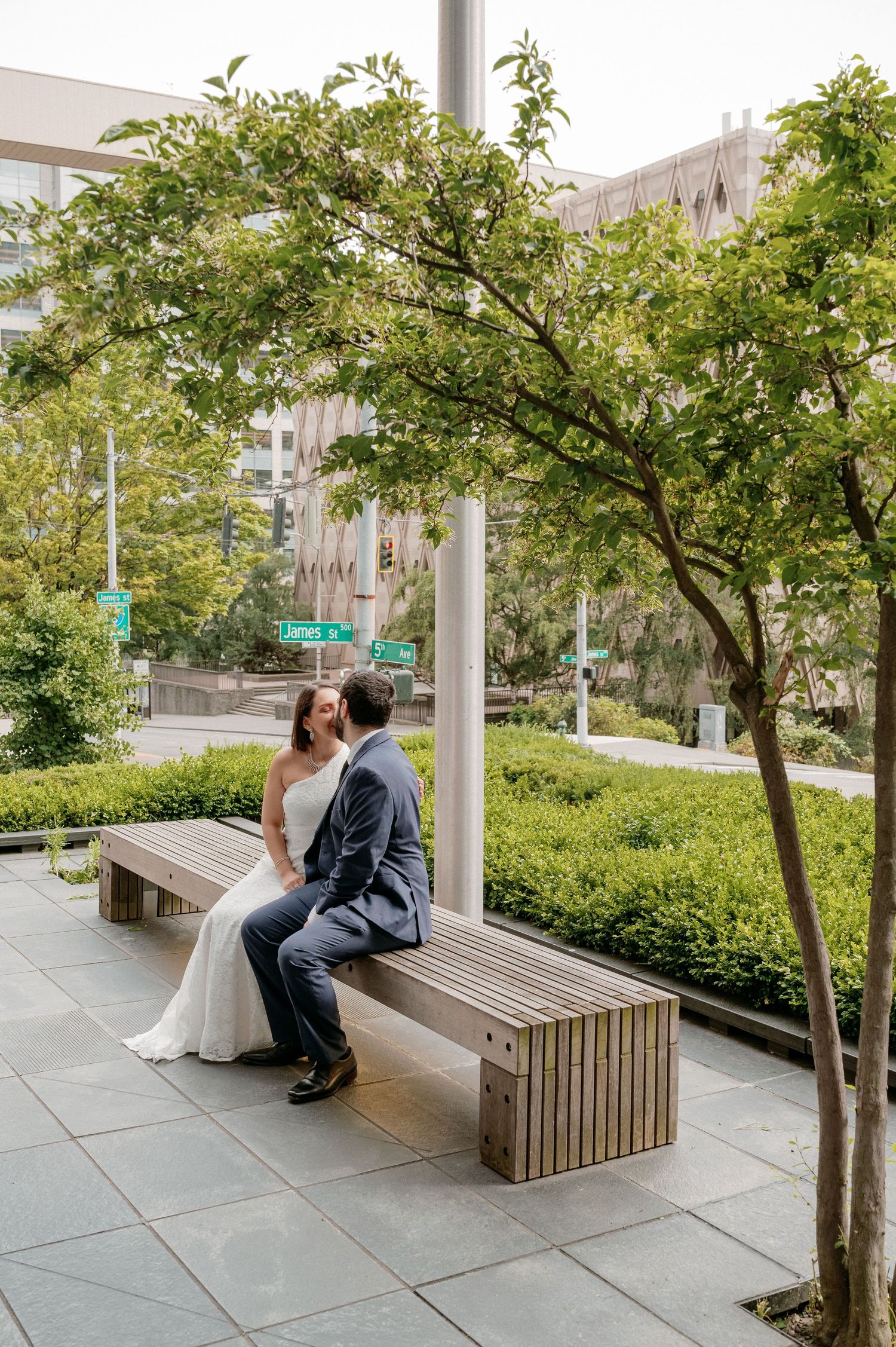 A couple in wedding attire sits together on a wooden bench beneath a green tree in an urban courtyard.