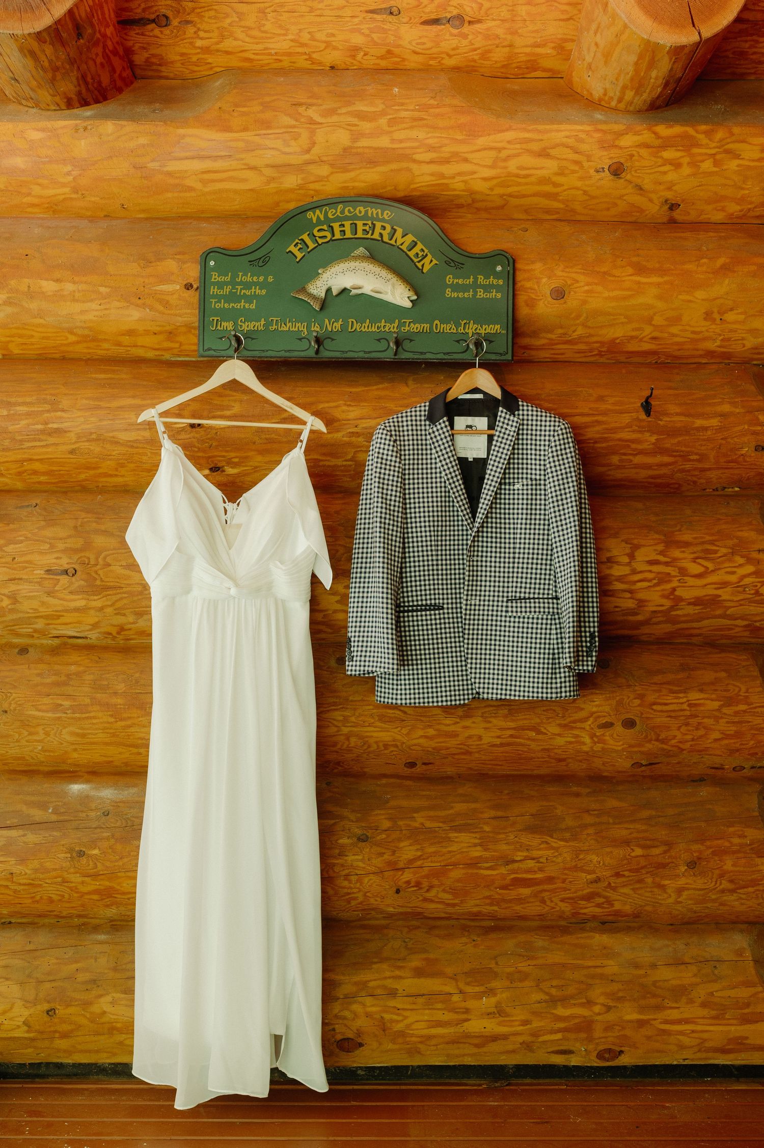 White wedding dress and plaid blazer hanging on rustic log cabin wall beneath a Welcome Fishermen sign.