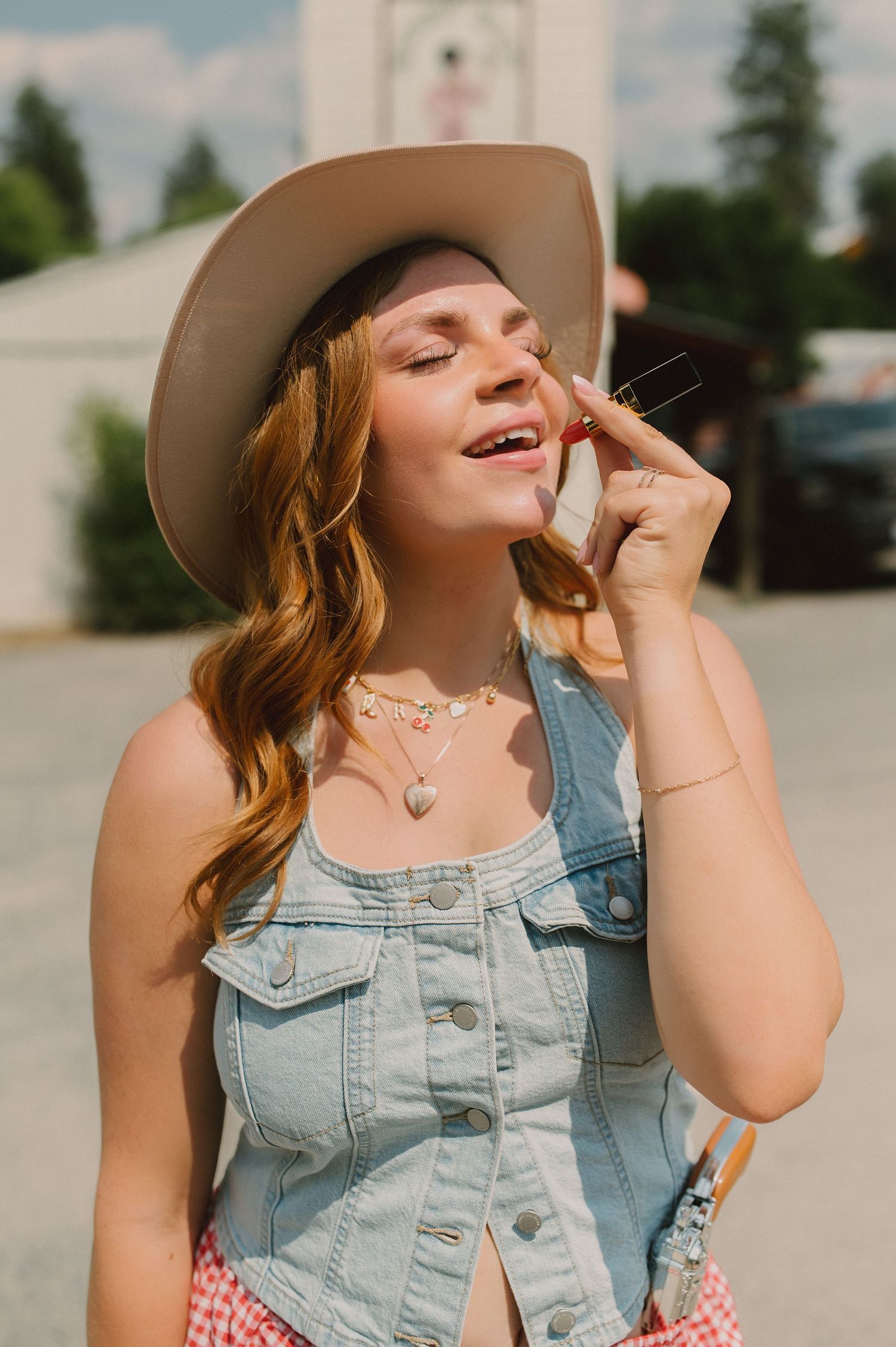 A person in a denim vest and beige hat smiles joyfully while wearing layered necklaces on a sunny day.
