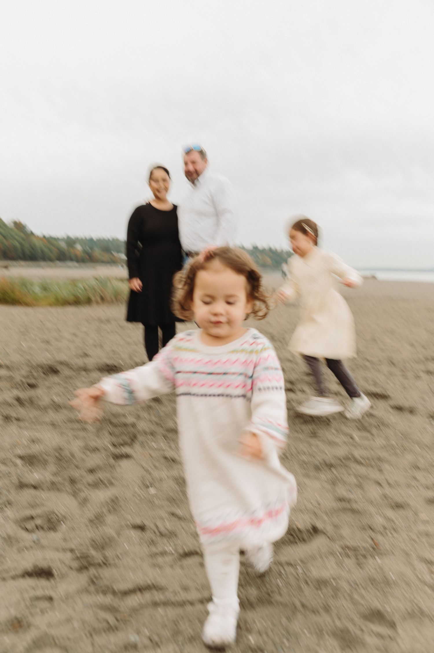A family enjoys a playful moment together on a cloudy beach with a small child running in the foreground.