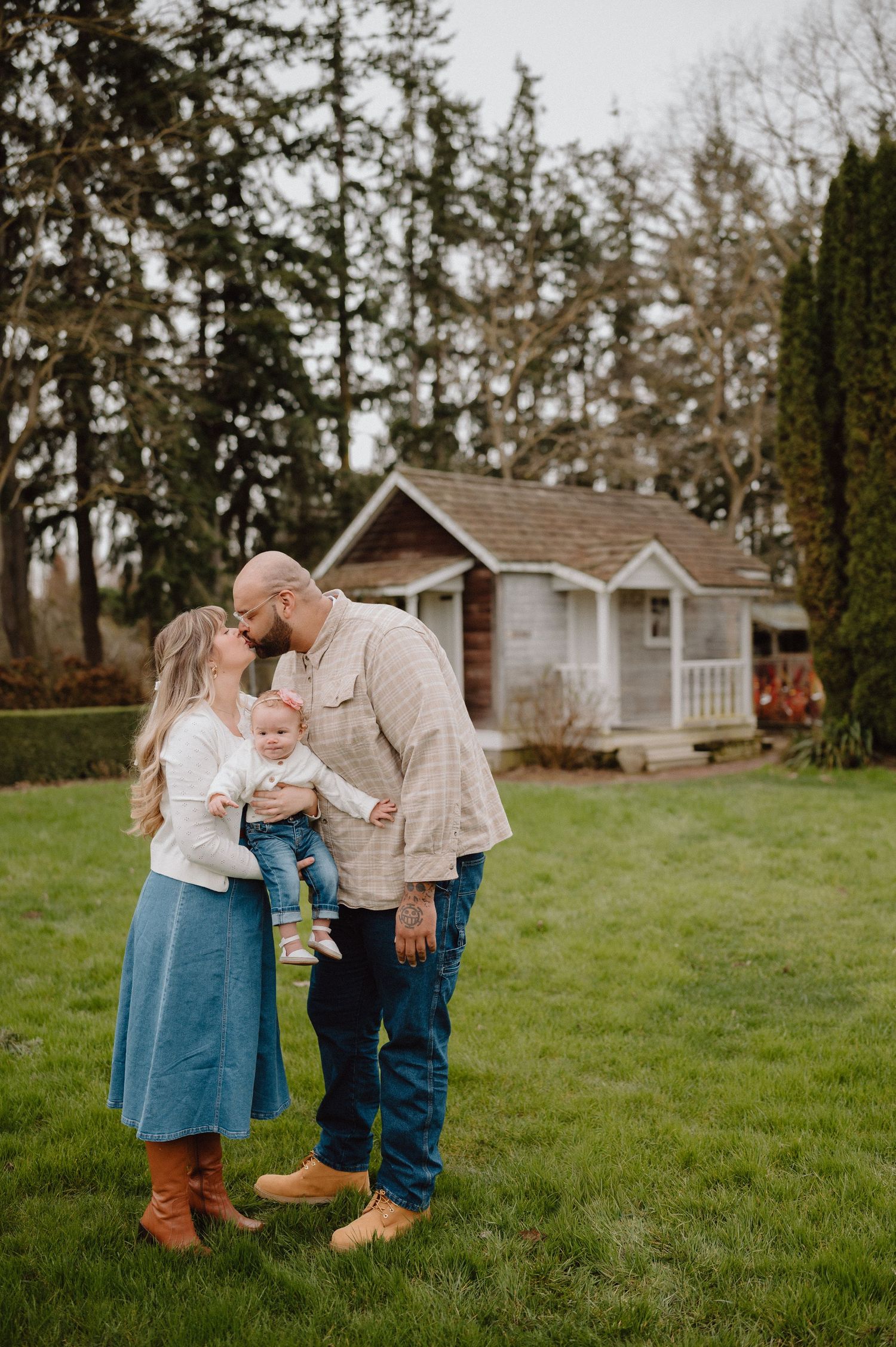 A couple shares a kiss while holding their baby in a lush green yard with a charming cottage and tall trees behind them.