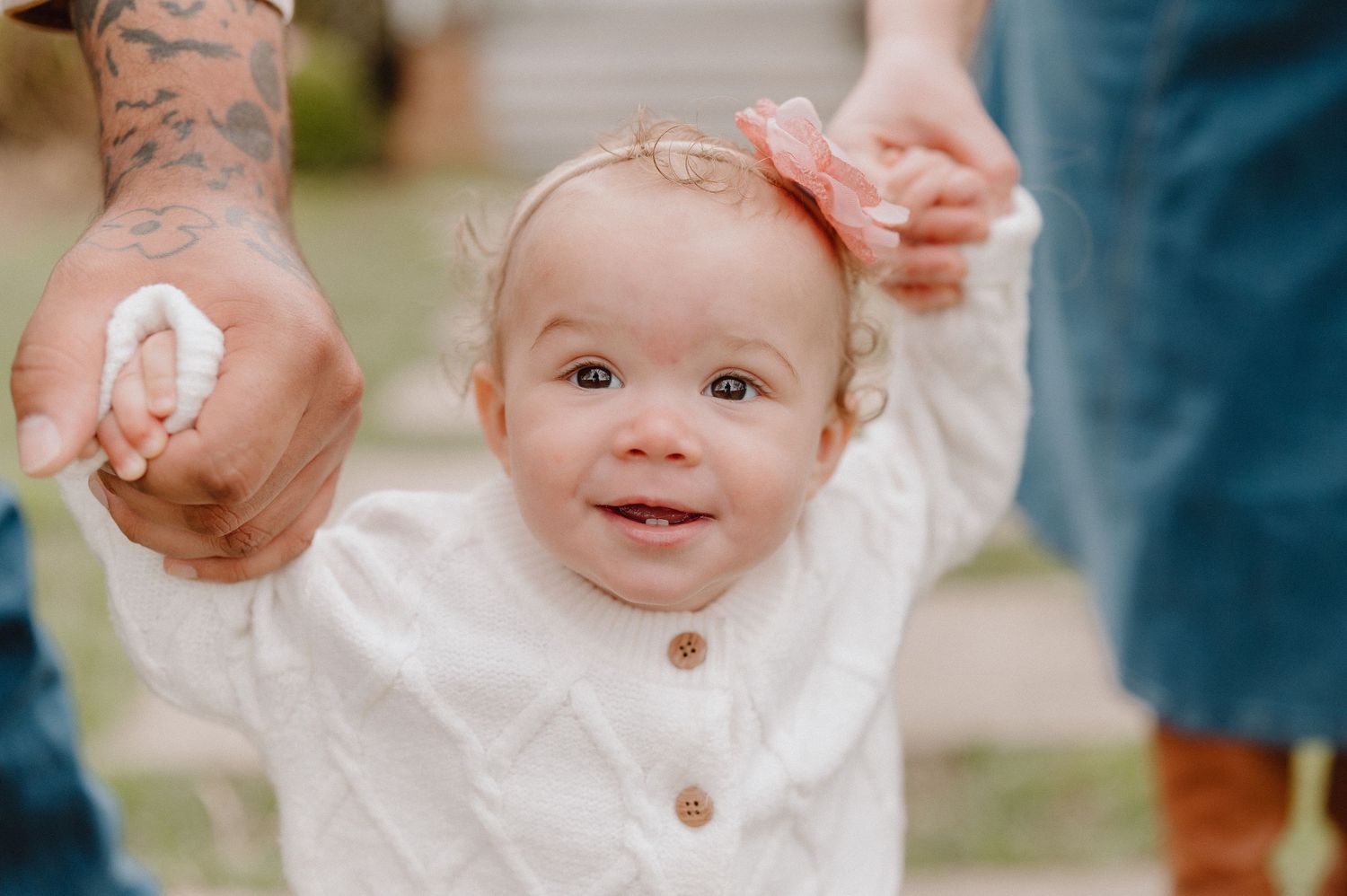 Smiling baby girl with pink bow headband holds adults hands while learning to walk outdoors.