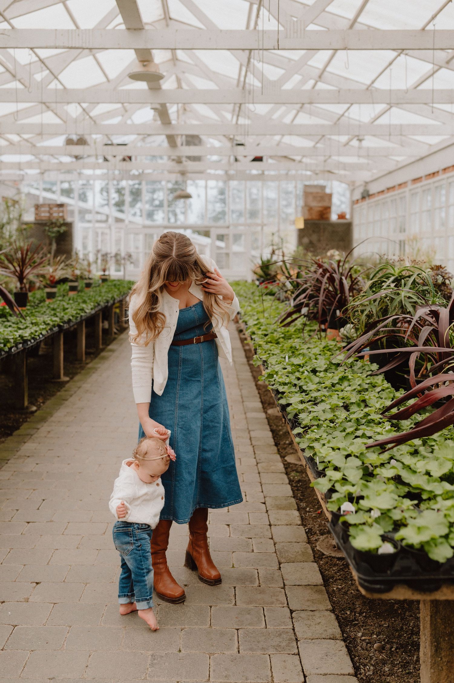 A mother and toddler walk through a lush greenhouse, surrounded by rows of green plants and colorful foliage.