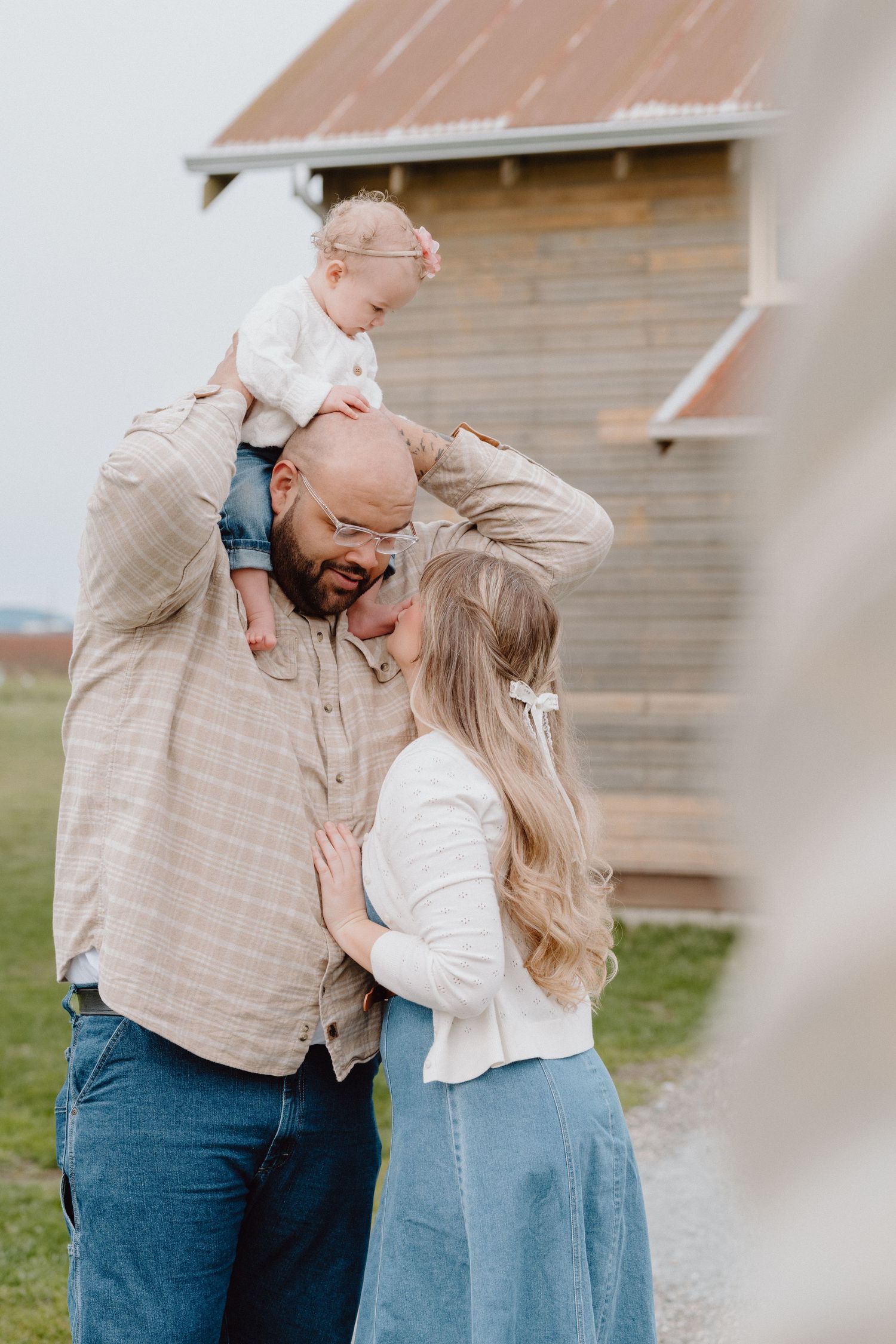 A happy family with a bald father, blonde mother, and baby on shoulders pose near a rustic barn outdoors.