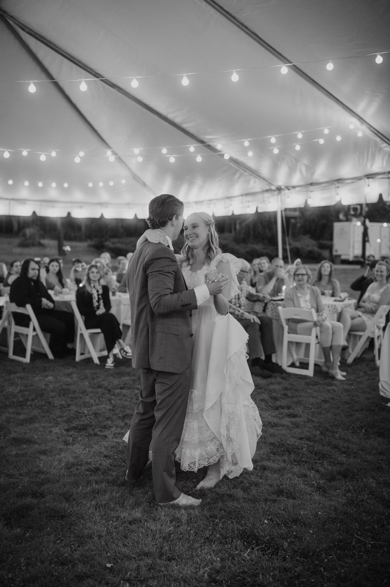 Bride and groom share first dance under string lights in a tent at an outdoor wedding reception.