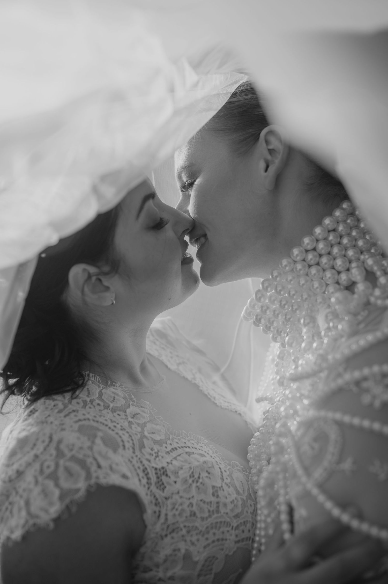 Intimate black and white portrait of two brides under a veil in lace wedding dresses.