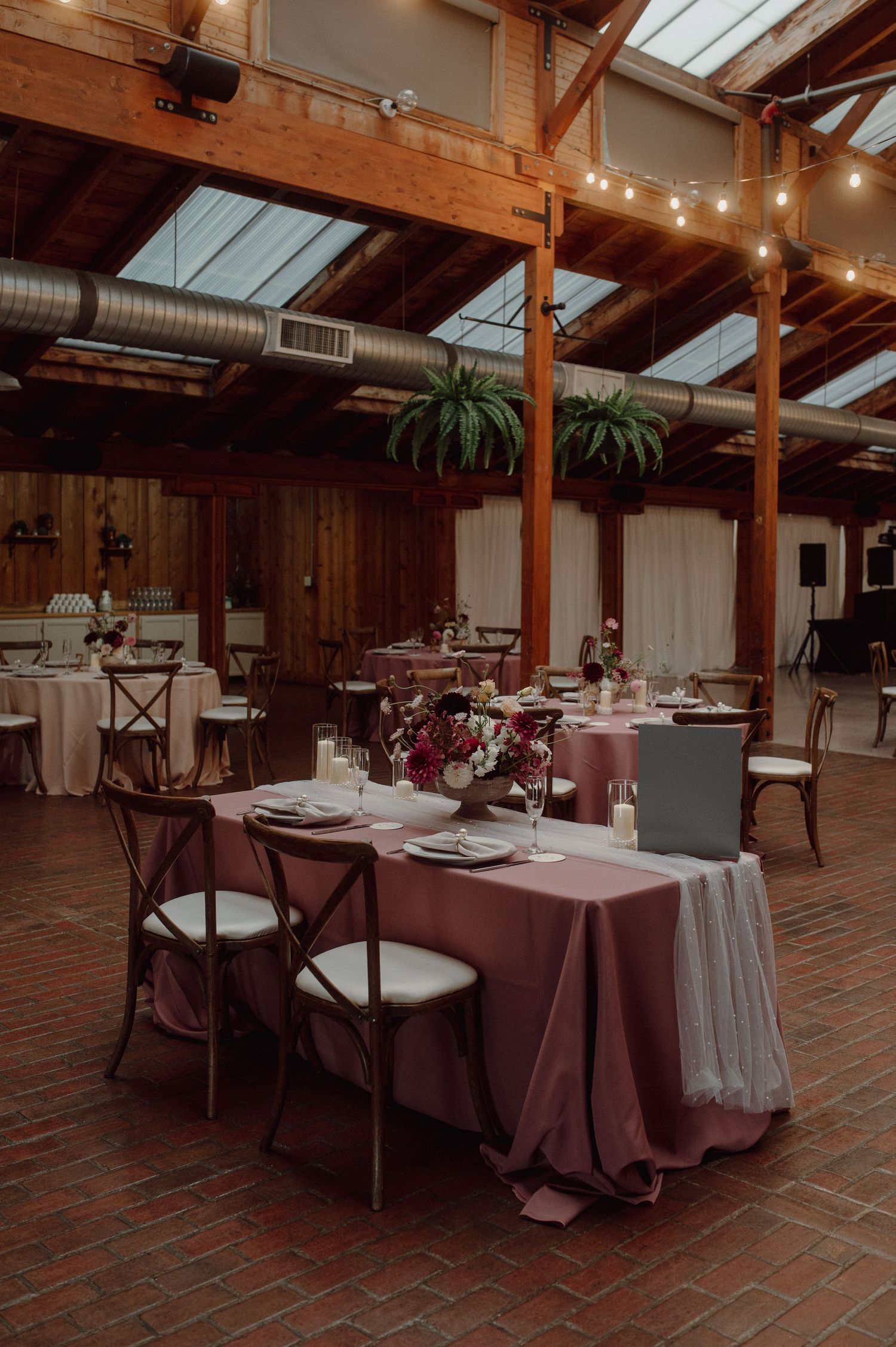 Rustic indoor wedding reception venue with exposed wooden beams, string lights, and elegant table settings on brick flooring.