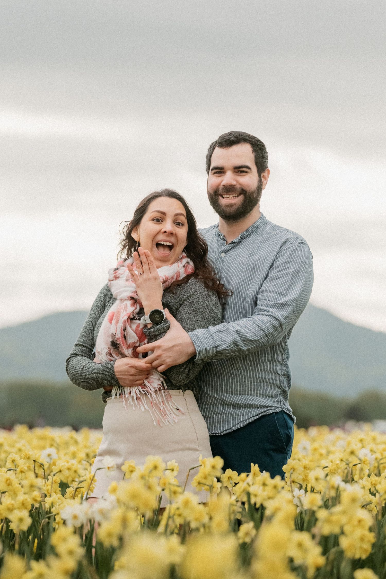 Happy couple embracing in field of yellow daffodils with mountains in background on overcast day.