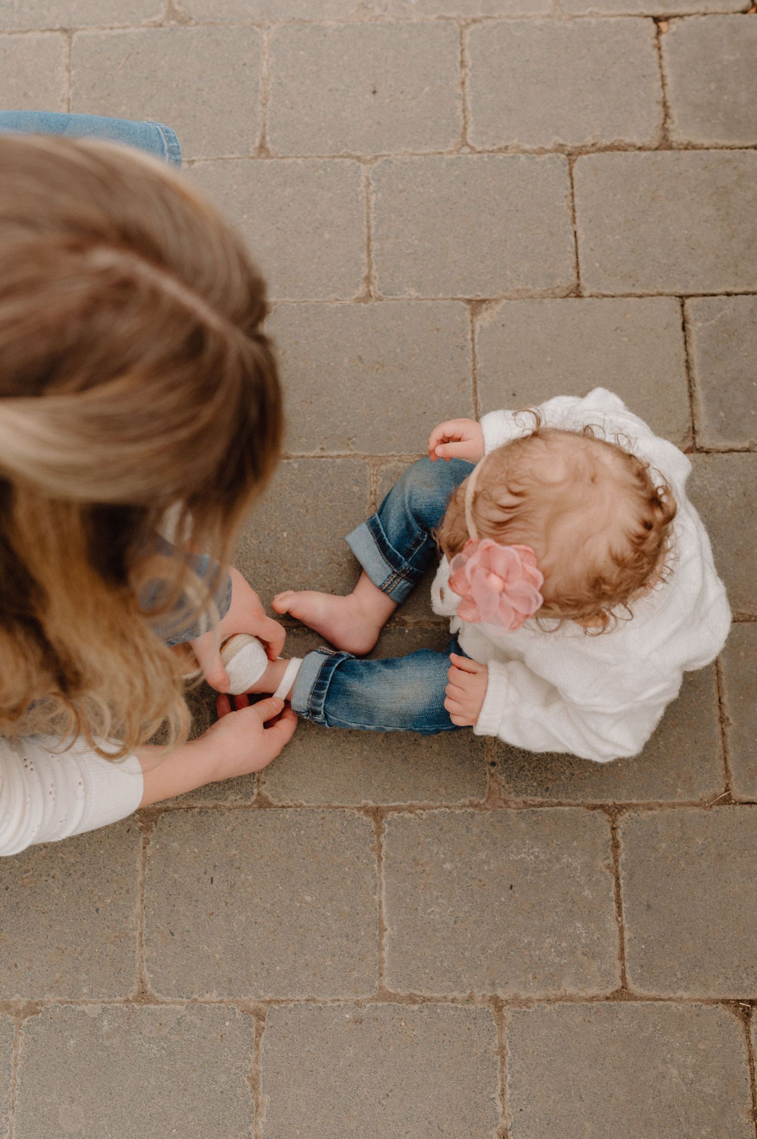 Overhead view of a mother helping her toddler daughter sit on a brick patio, child wearing jeans and white bow.
