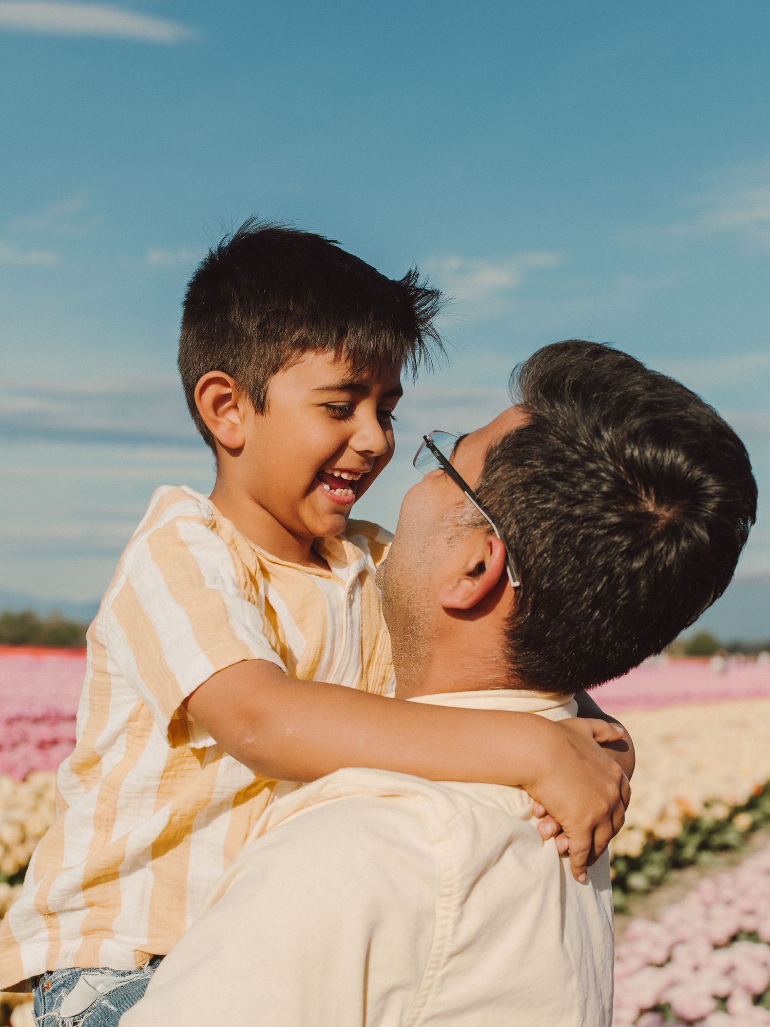 Playful father and child share a joyful embrace outdoors with blooming pink flowers in the background.