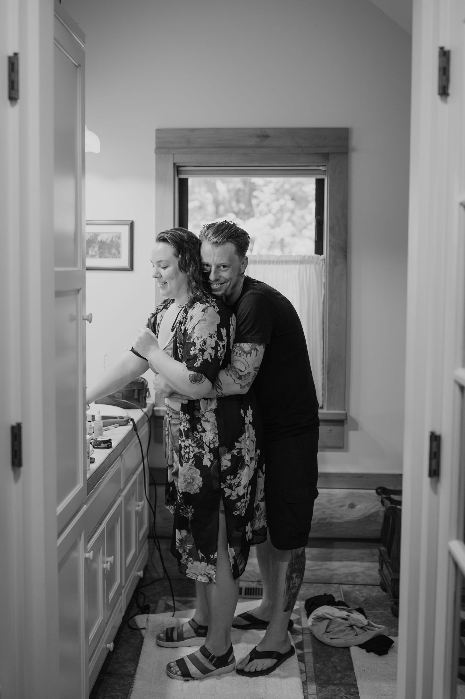 Black and white photo of a couple embracing in a kitchen, man hugging woman from behind as she stands at the counter.