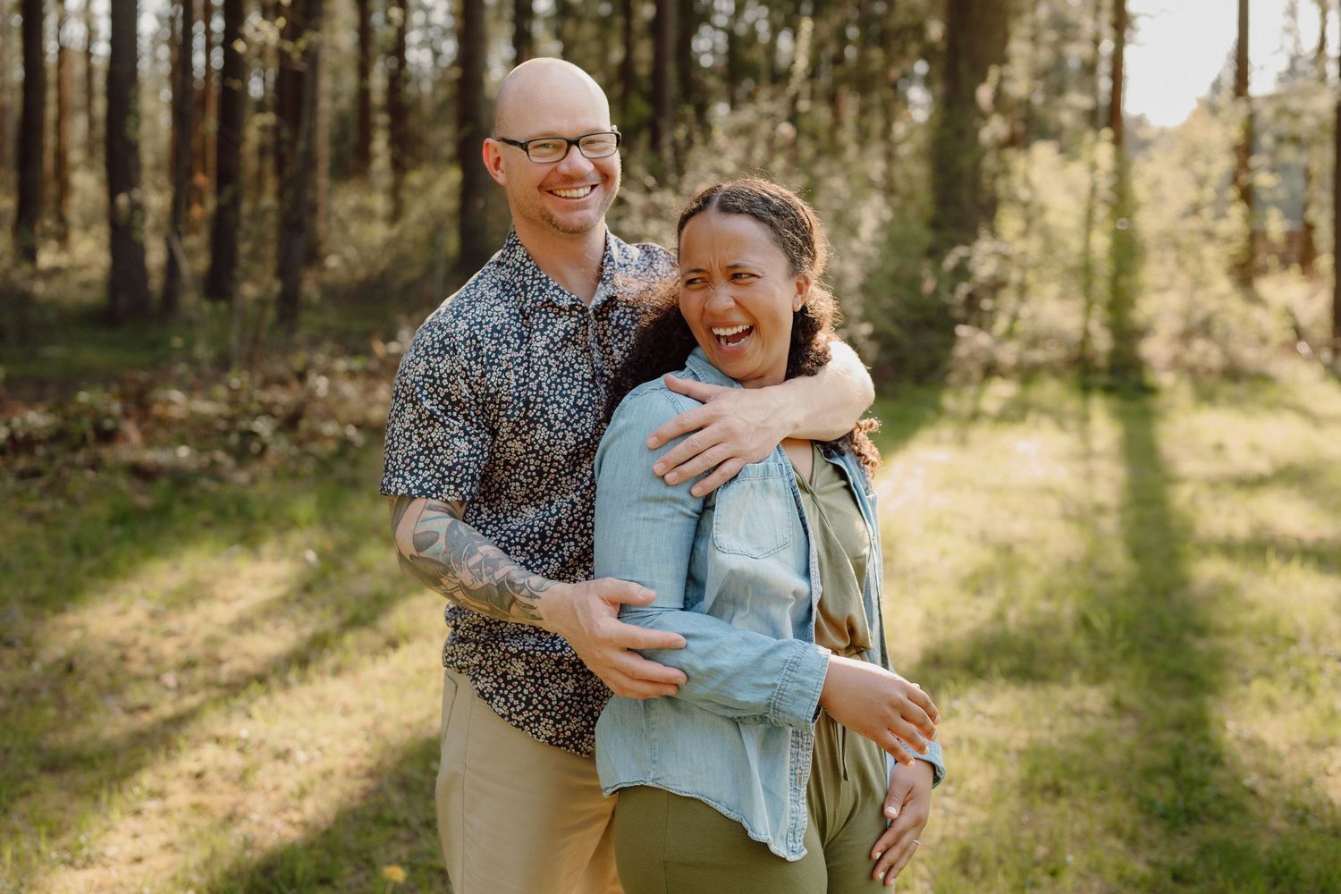 Two people embrace warmly in a sun-dappled forest setting wearing casual attire.