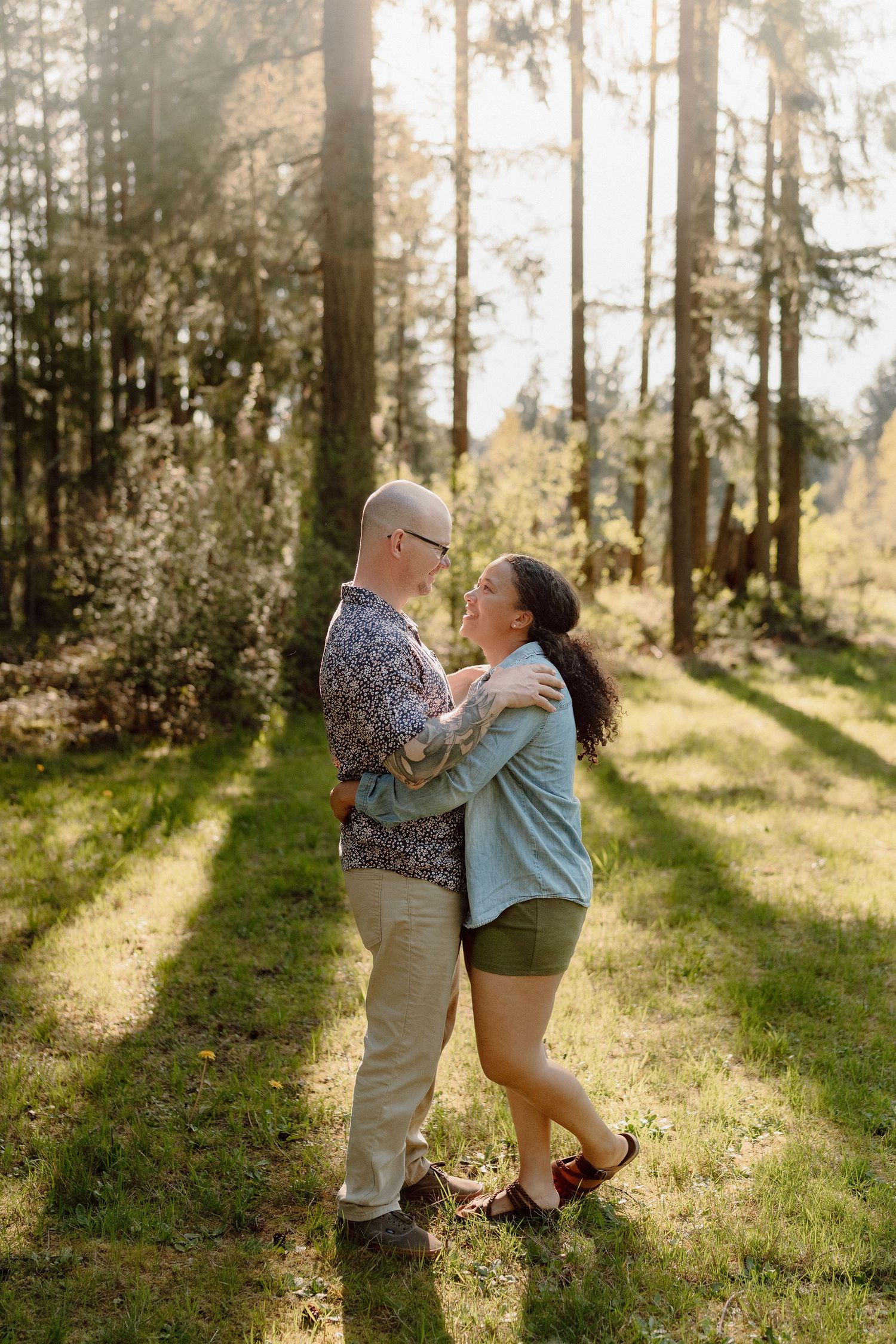 A couple shares a romantic moment in a sunlit forest clearing, embracing while surrounded by tall evergreen trees.
