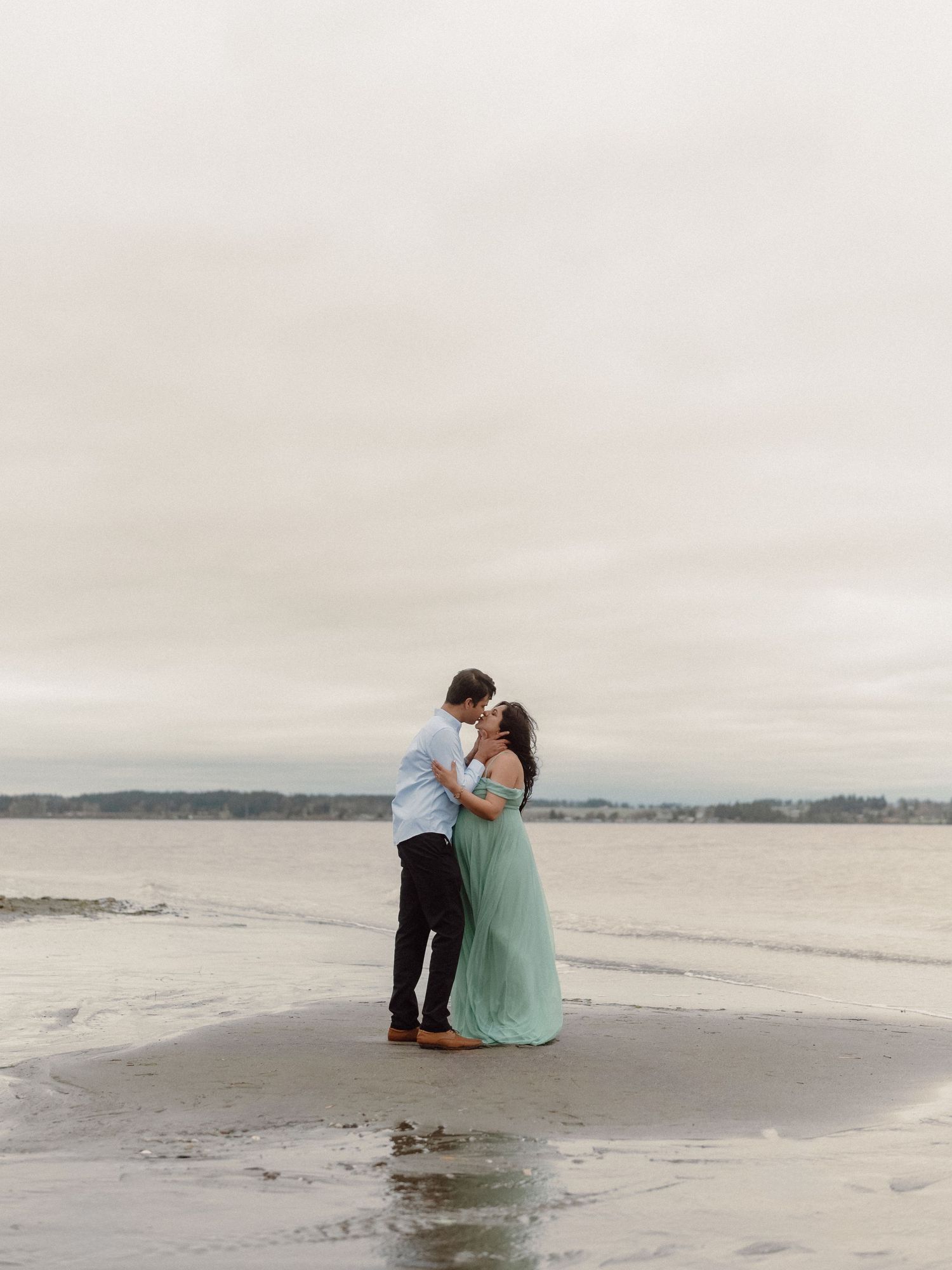 A couple shares a romantic moment on a misty beach shoreline in coordinating mint and white outfits.