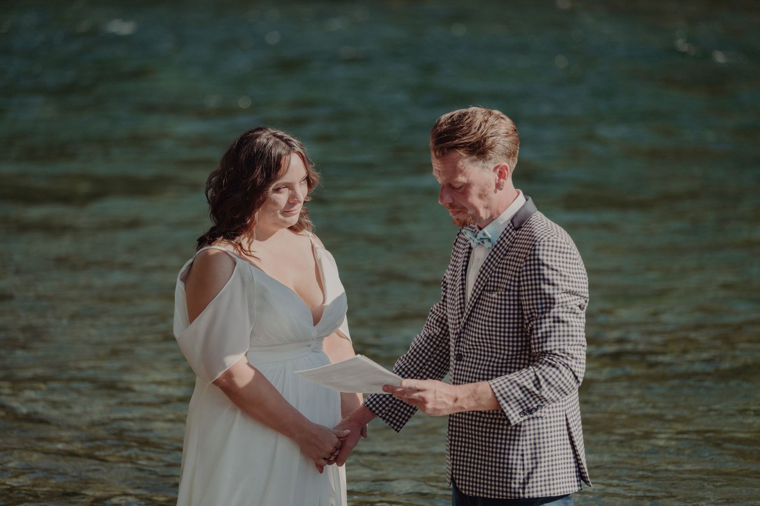 Couple exchanging vows by a river, bride in white gown and groom in checkered blazer reading from paper.