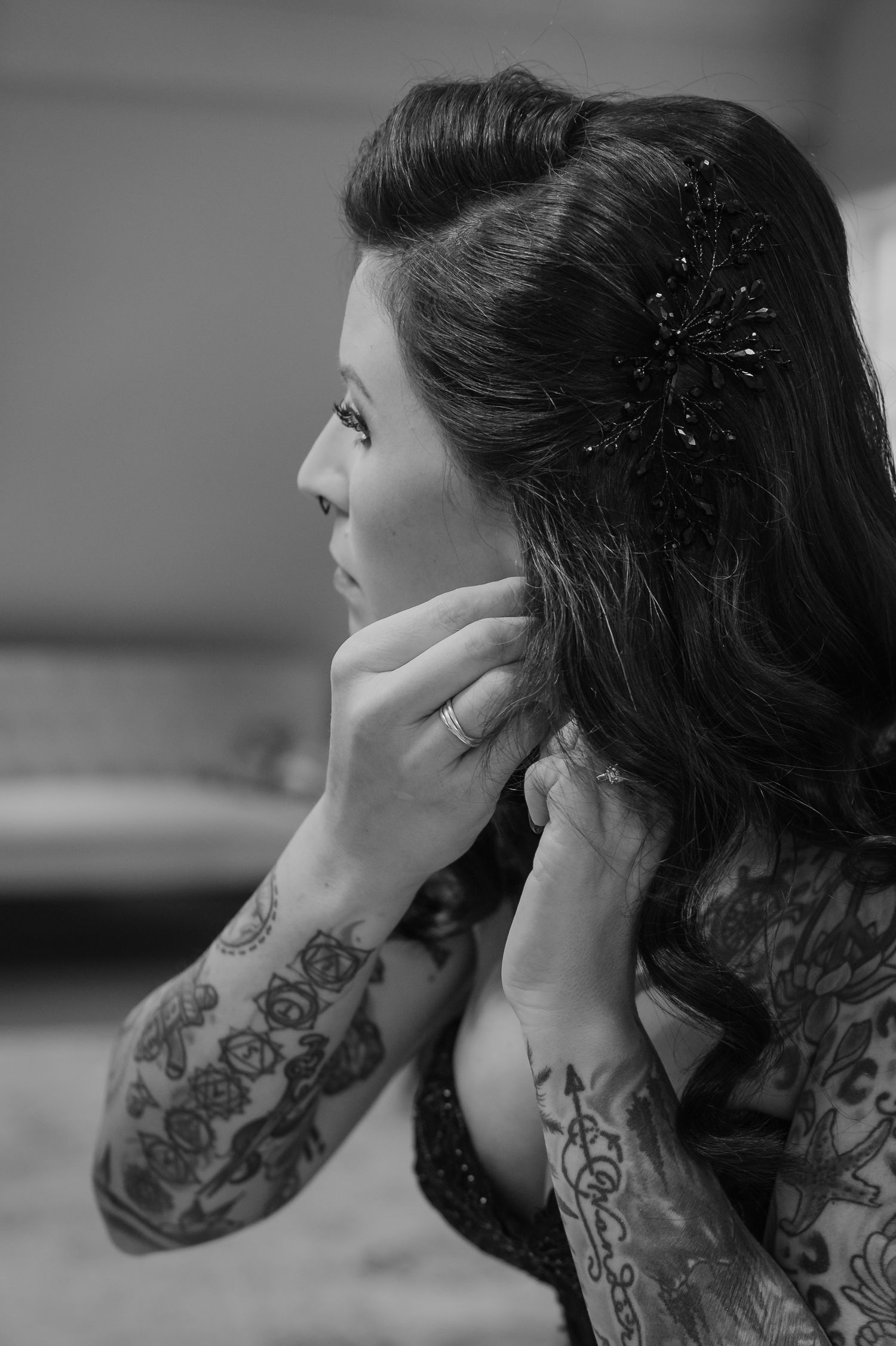 Black and white photo of a tattooed woman adjusting her earring, wearing an elegant updo with a decorative hair accessory.