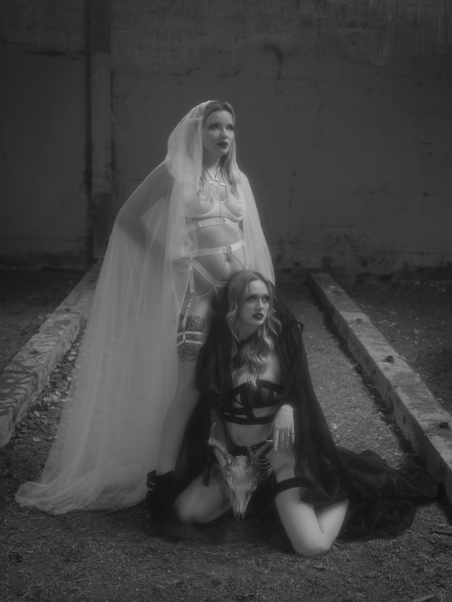 A person in gothic bridal attire wearing white veil and black lingerie poses dramatically on railroad tracks in black and white.