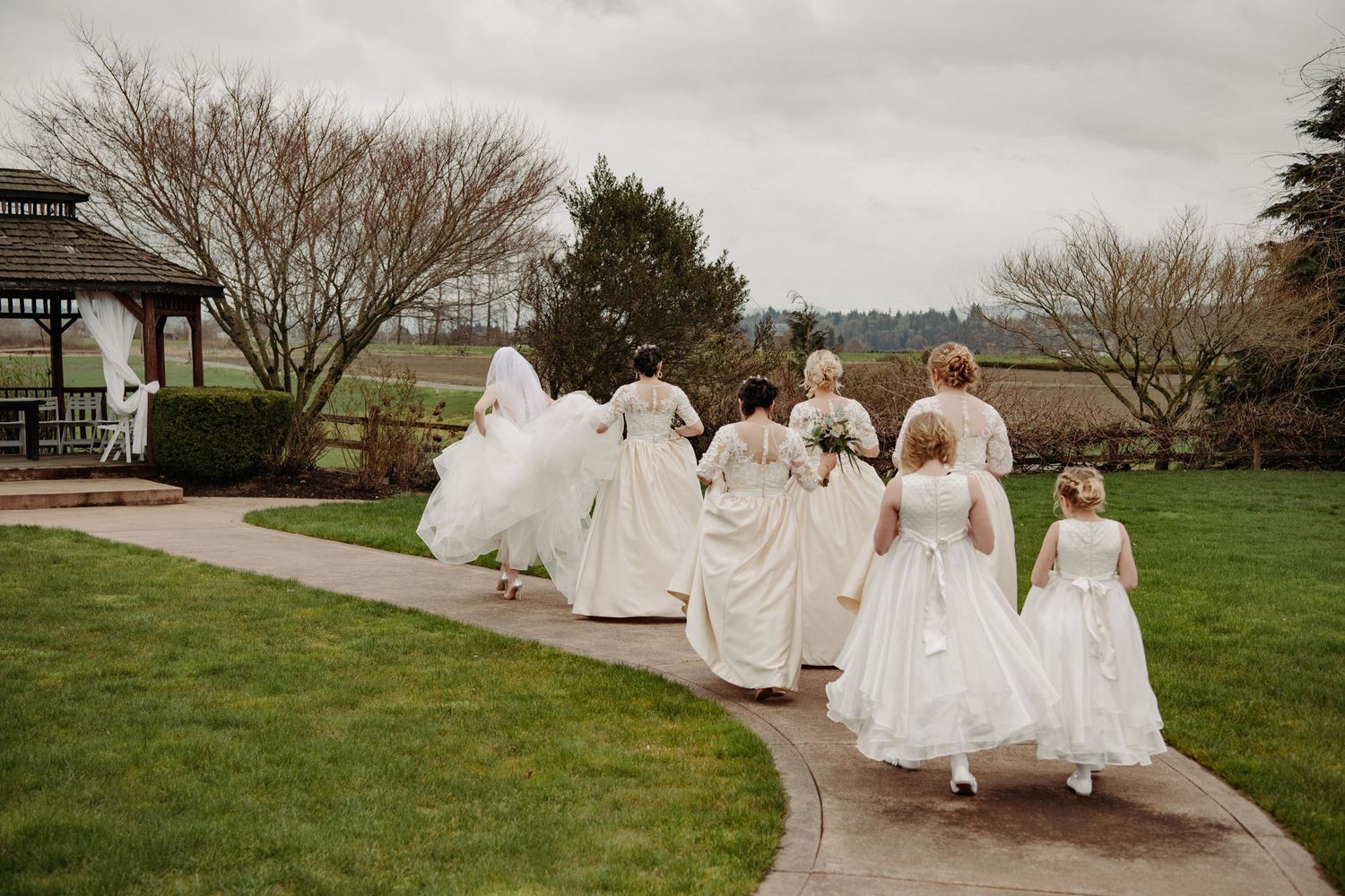 A line of bridesmaids in white dresses walking along a garden path.