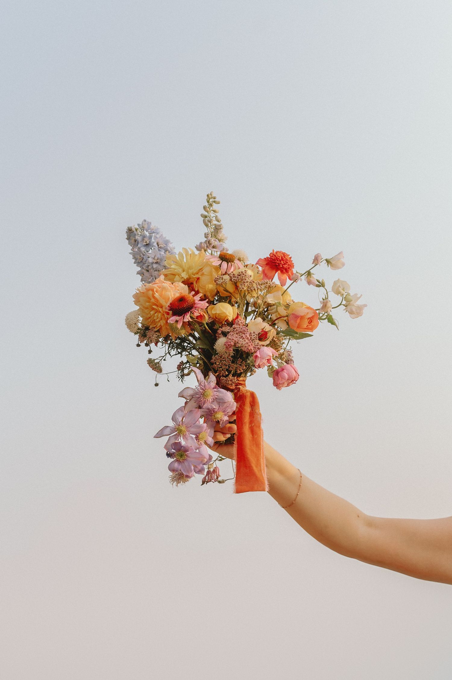 Hand holding a vibrant bouquet of mixed flowers in peach, pink and orange against a light background.