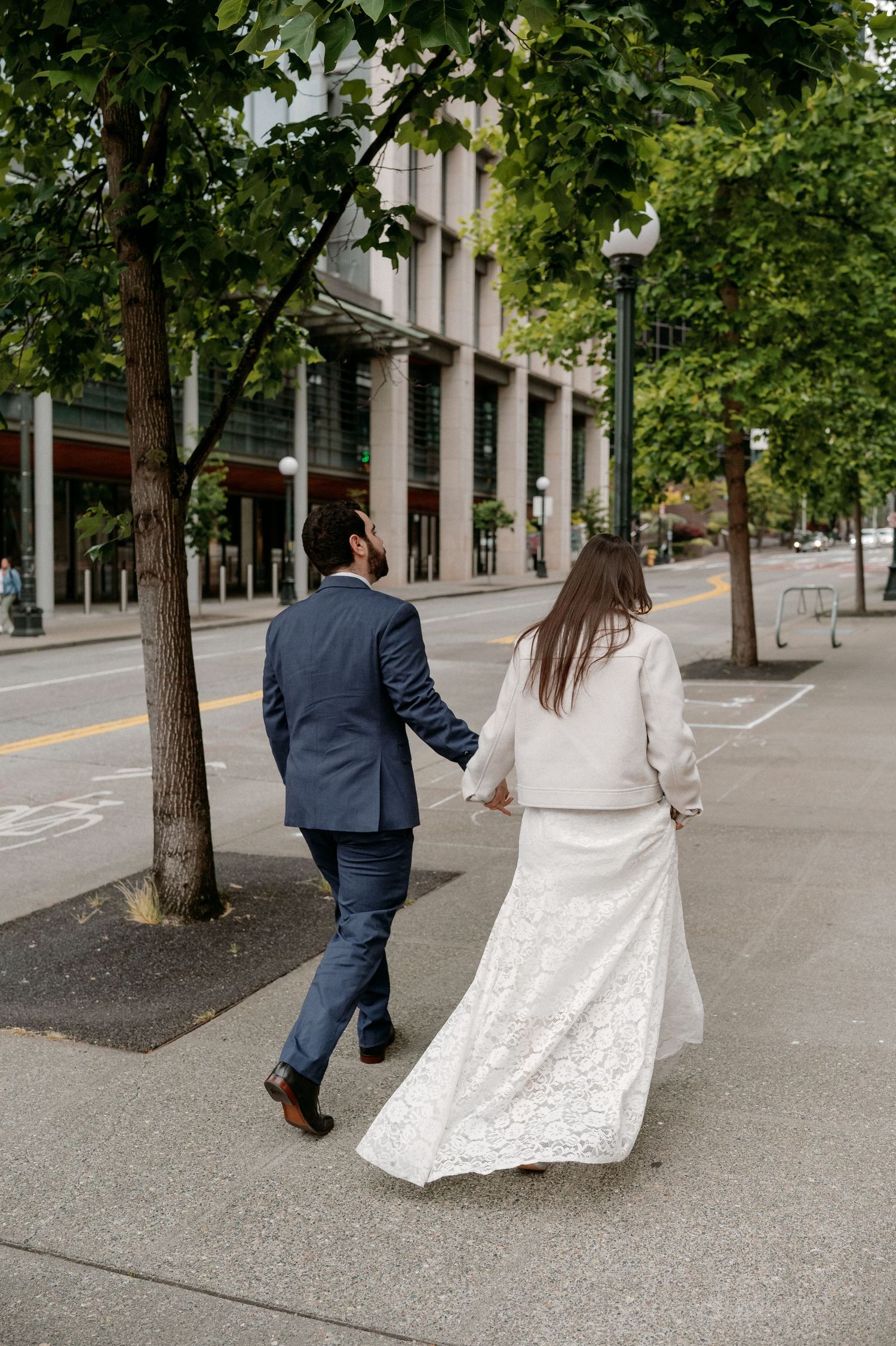 Couple in wedding attire walking together on a tree-lined city sidewalk with urban buildings in the background.