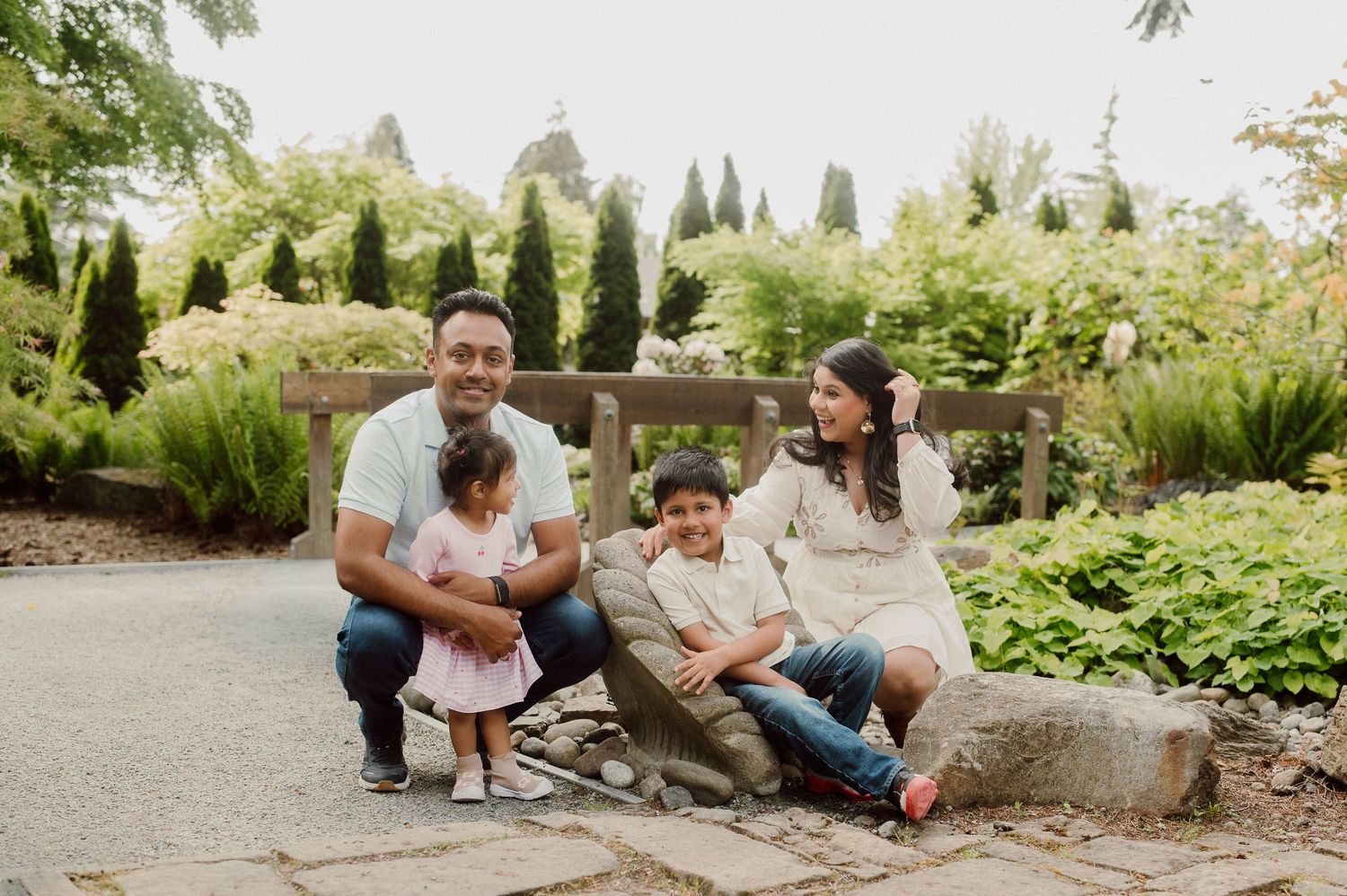 Family of four smiling outdoors in a lush garden, parents with two young children seated on a stone bench.