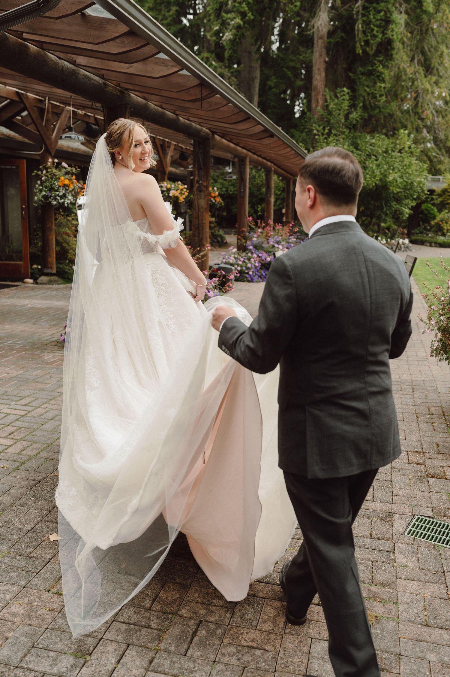 A couple in formal wedding attire walking together on a stone pathway under a wooden overhang.