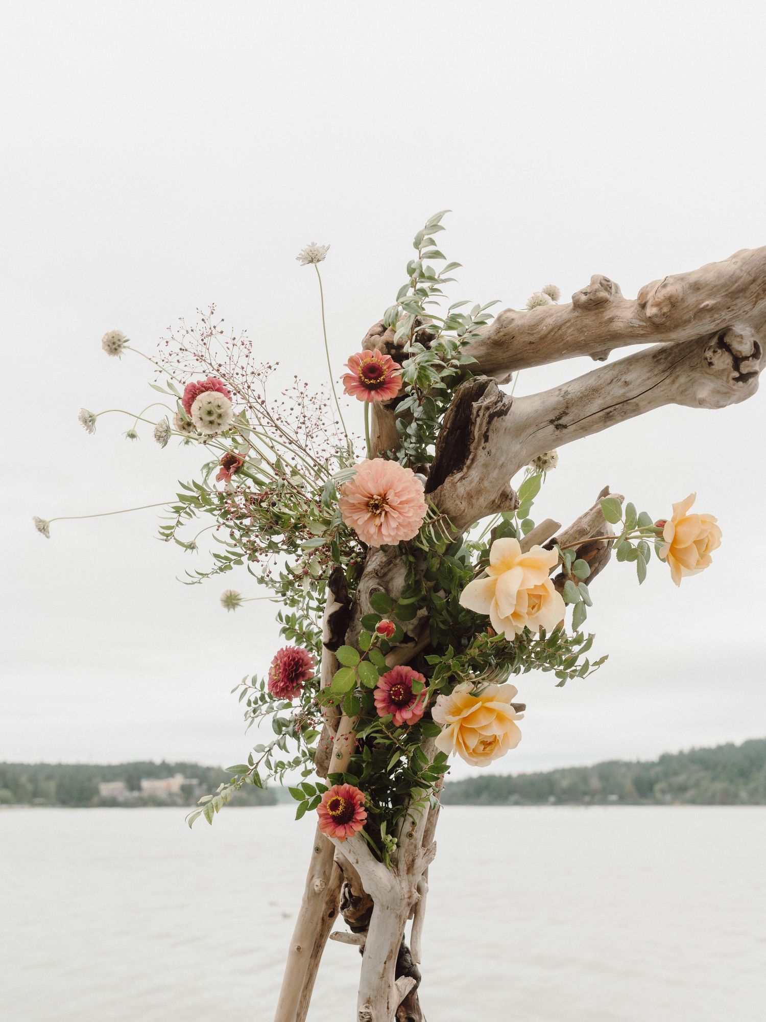 Rustic driftwood arch decorated with peach and burgundy flowers against a serene waterfront backdrop.