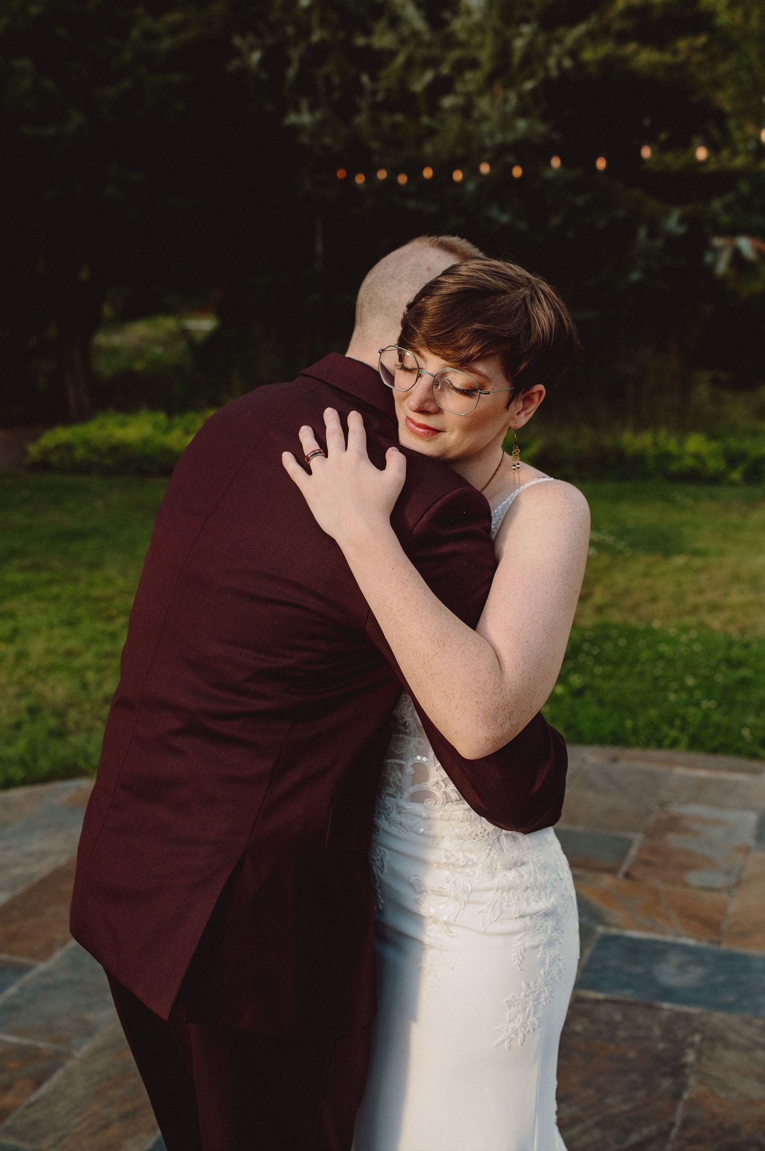 Couple sharing a tender embrace on their wedding day on a stone patio with string lights.