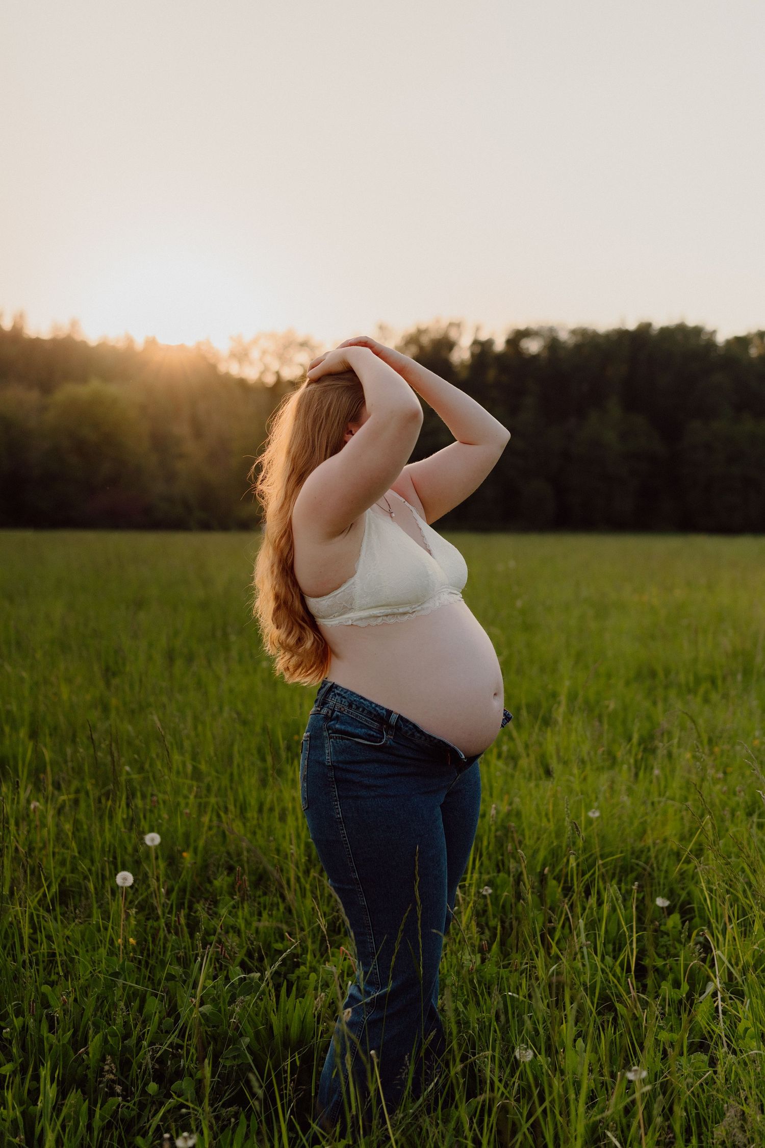 Expecting mother in white tank top poses in a green field at sunset.