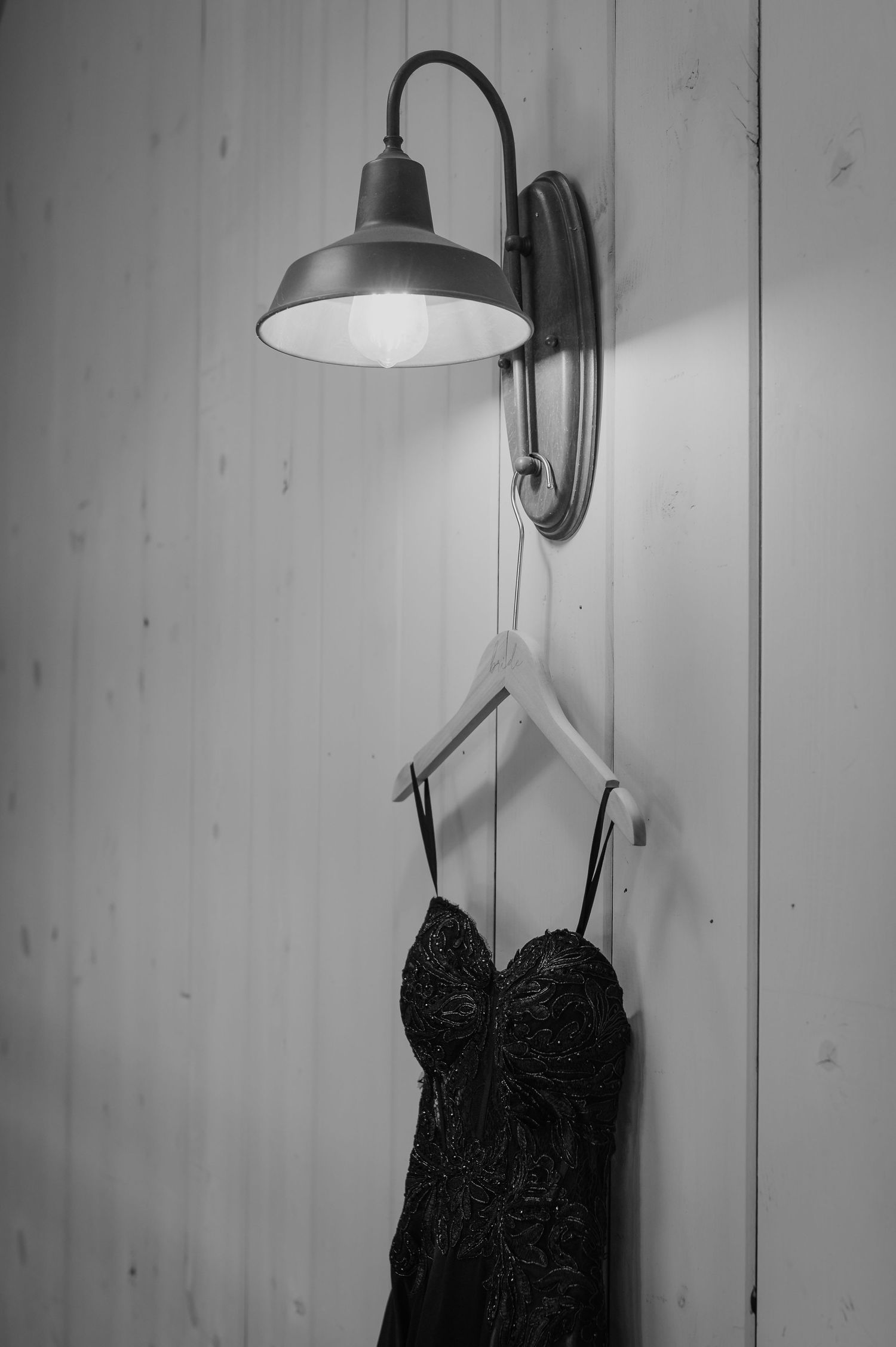 A black lace dress hangs on a wooden hanger beneath a wall-mounted industrial lamp in black and white.