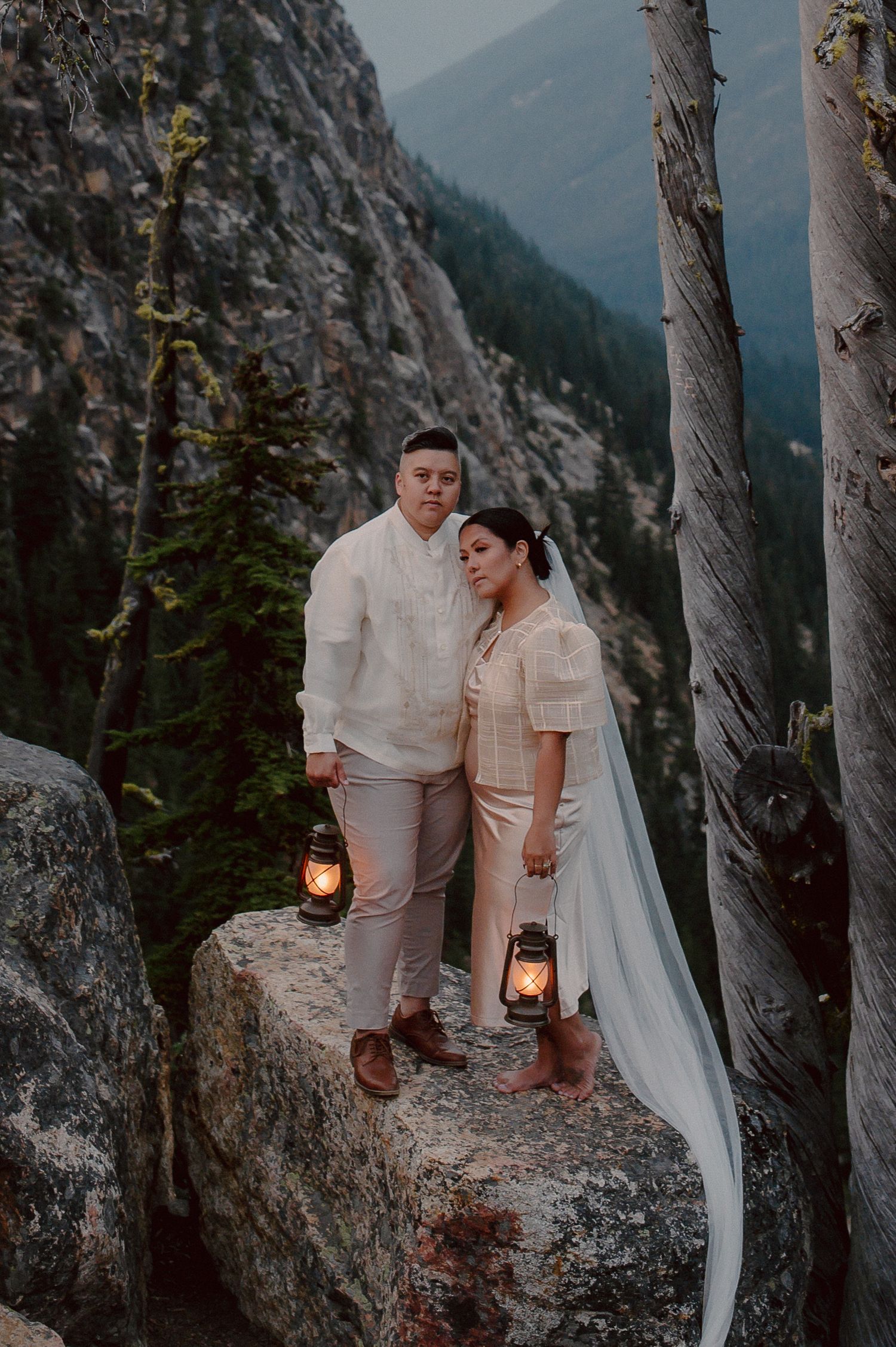A couple poses with lanterns on a rocky mountain ledge at dusk.