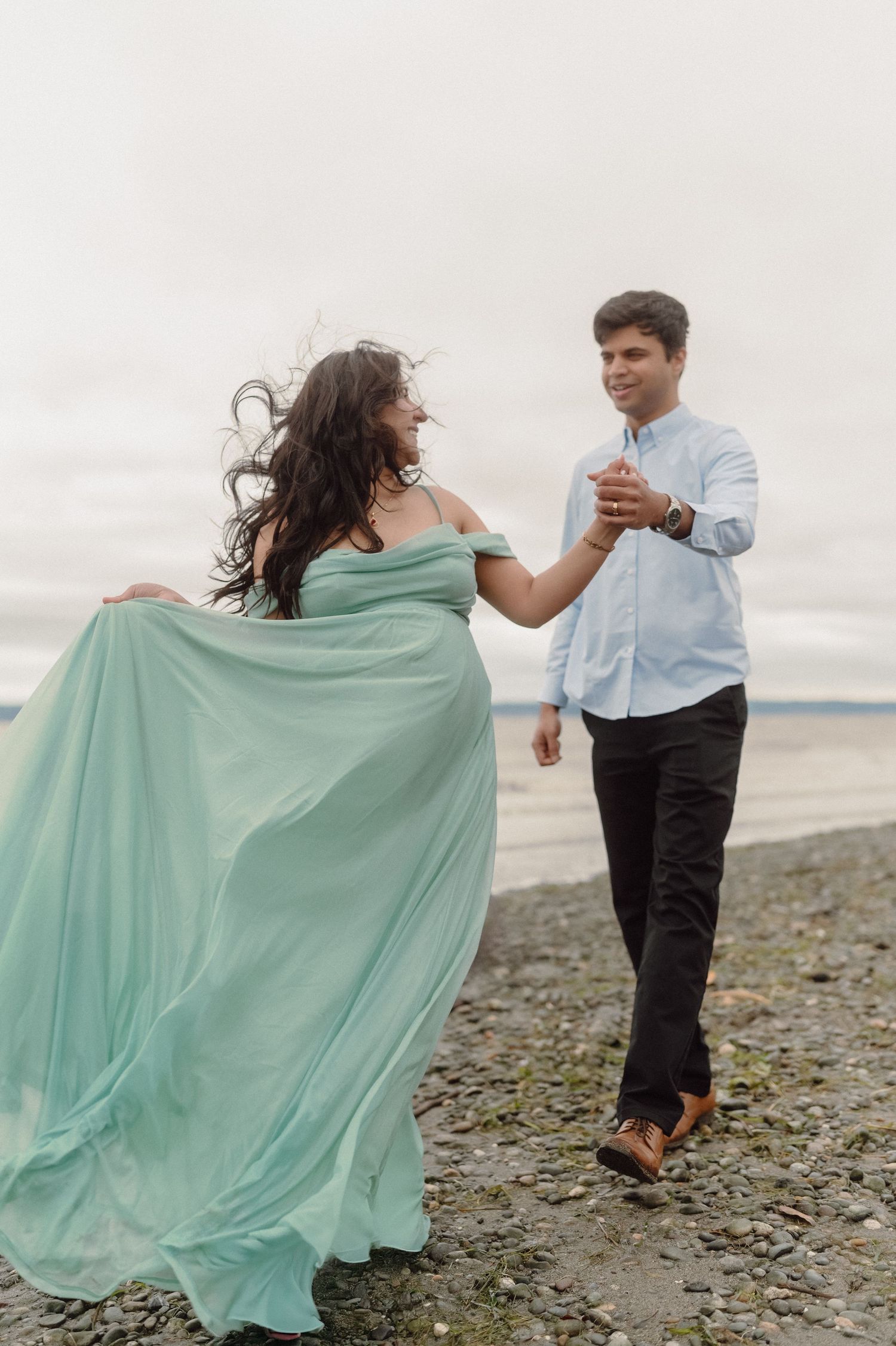 A couple dances playfully on a rocky beach, with the woman's mint green dress flowing in the wind against a cloudy sky.