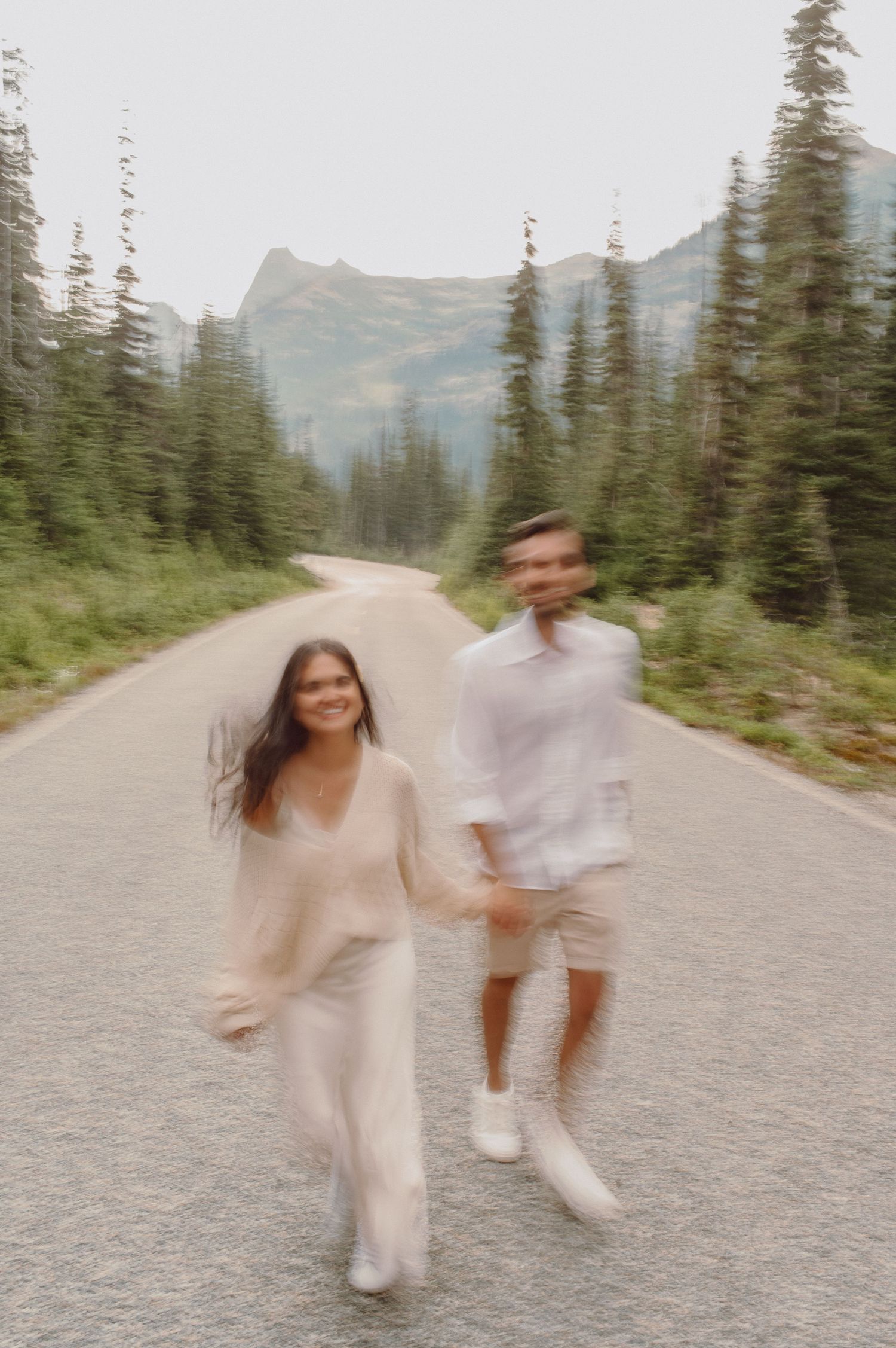 Two people dancing and twirling on a mountain road surrounded by evergreen trees and mountain peaks in the distance.