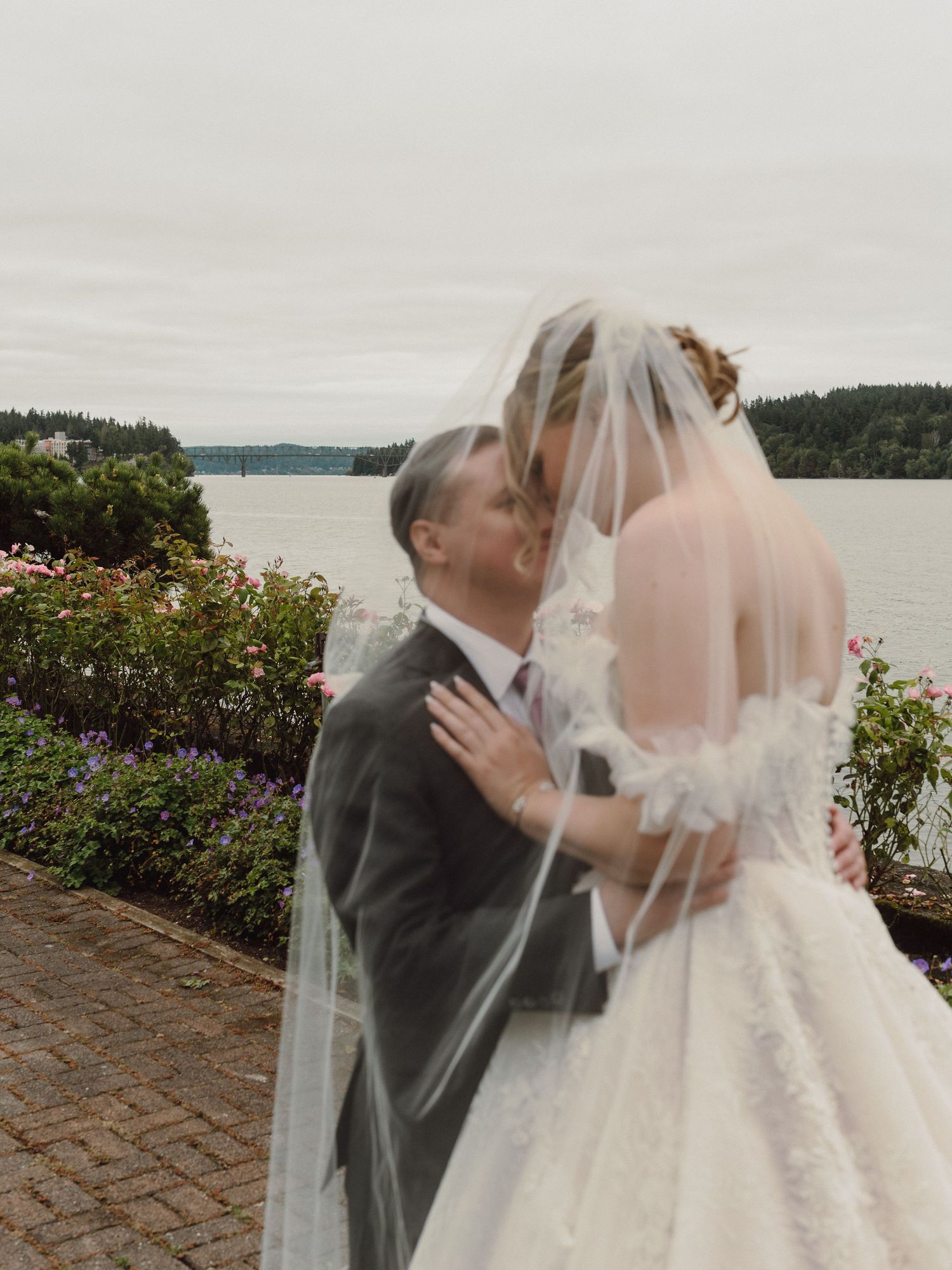 A romantic wedding couple shares a tender kiss on a brick patio overlooking a scenic waterfront view.