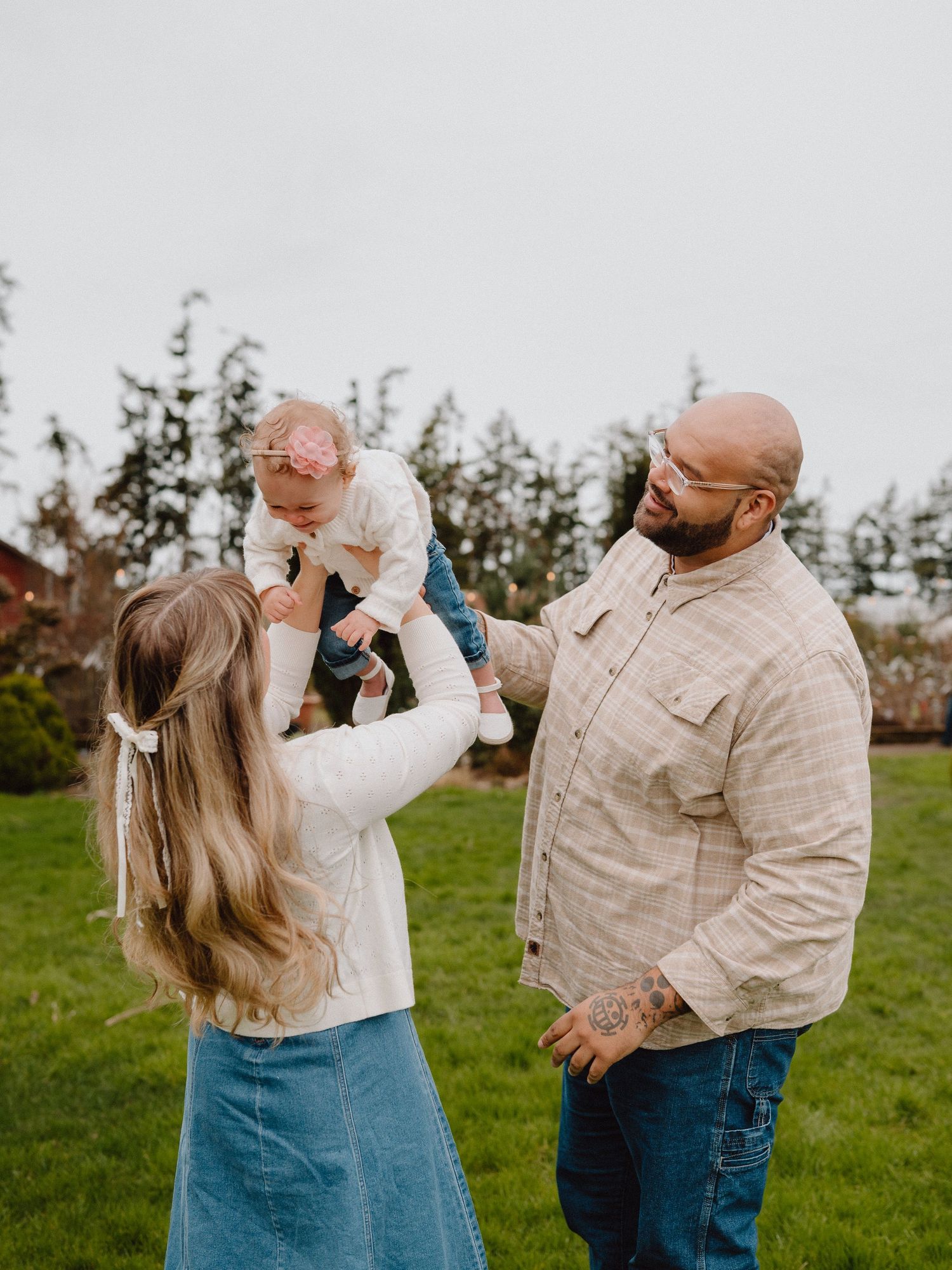 A young family with a bald tattooed dad and long-haired mom lifting their baby girl outdoors on a green lawn.