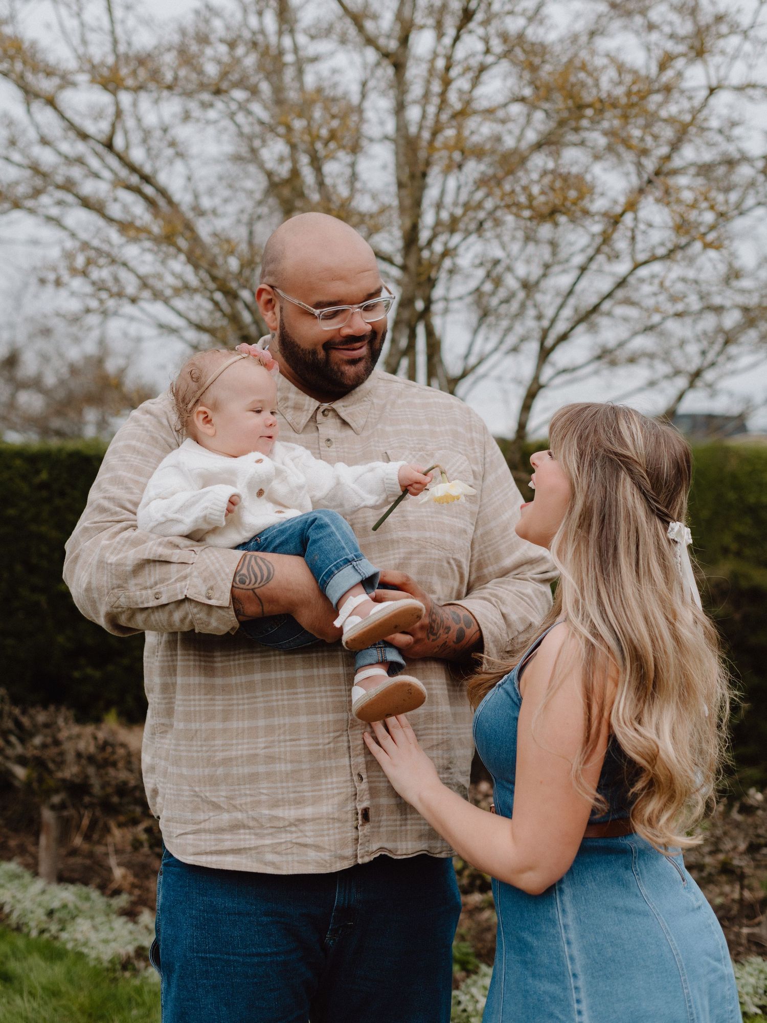 A bald man with glasses holds a baby while a blonde woman in a blue dress smiles at them outdoors.