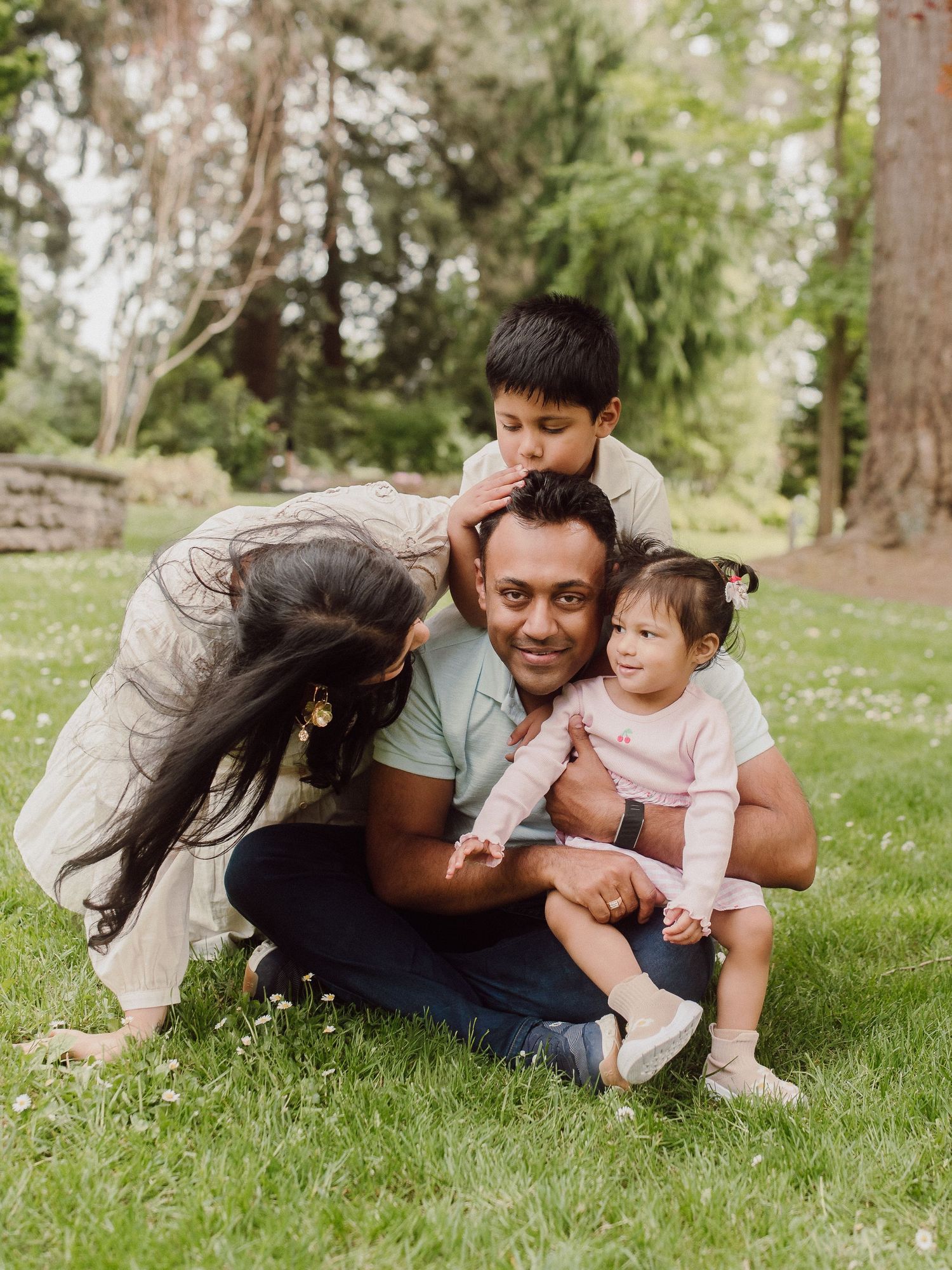A happy South Asian family of four poses together on green grass in a park, with two young children hugging their parents.