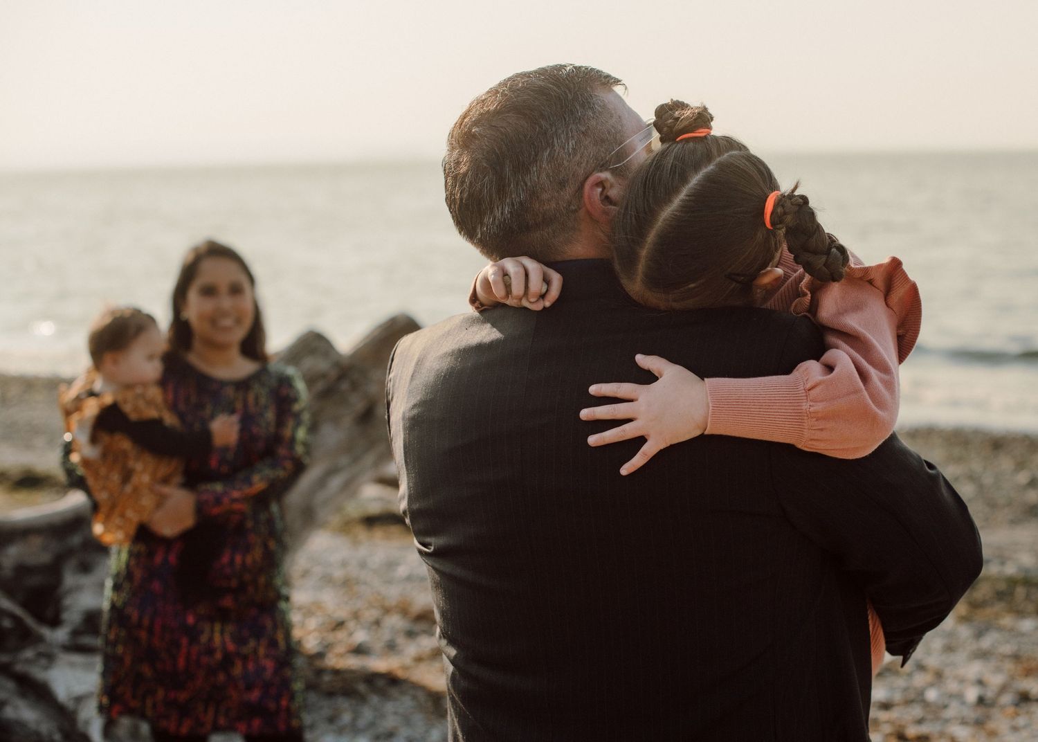 Family embraces on a rocky beach at sunset while someone photographs the tender moment in warm lighting.