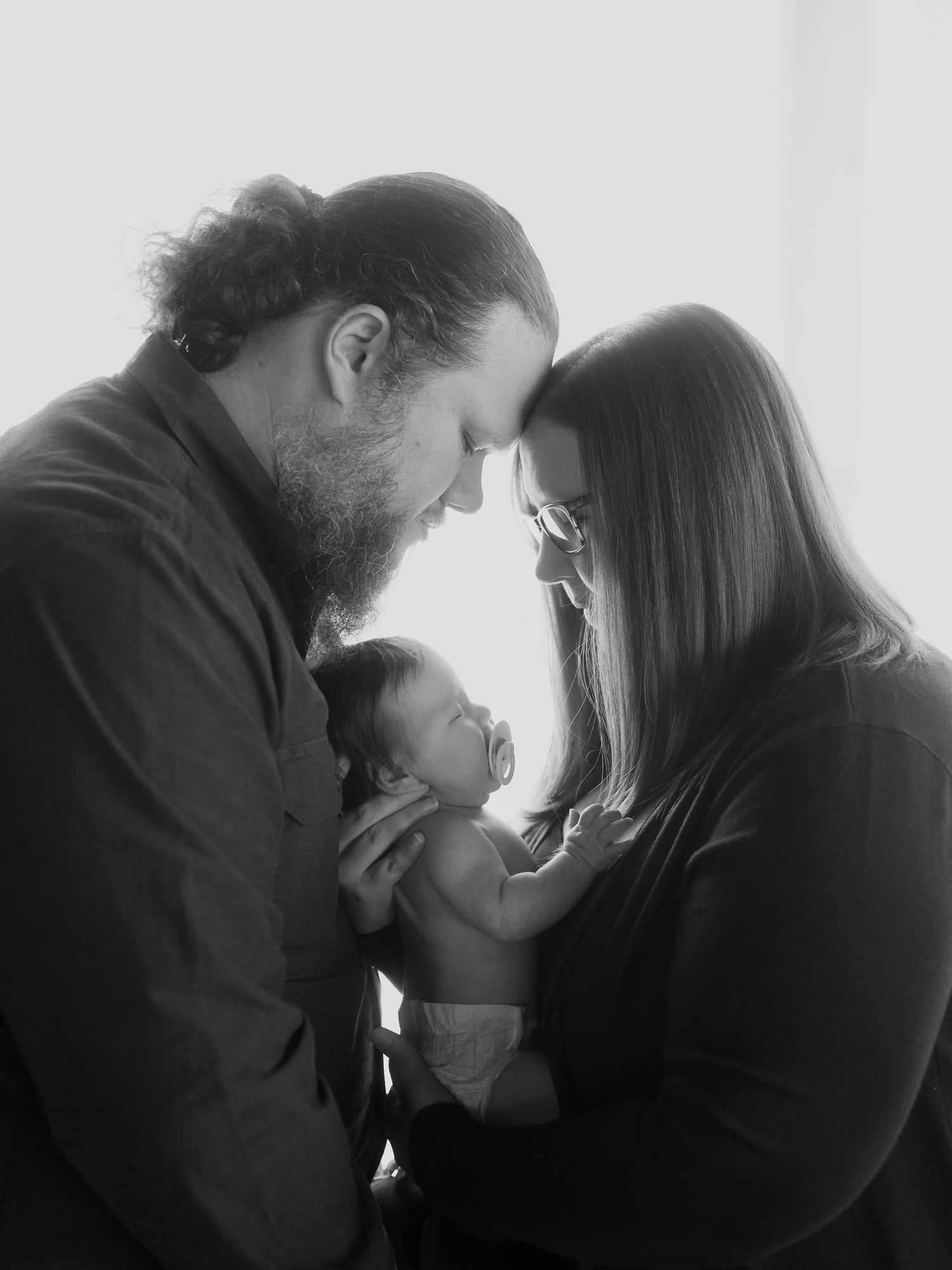 Black and white silhouette photo of two people with a newborn baby nestled between them in tender moment backlit by window.