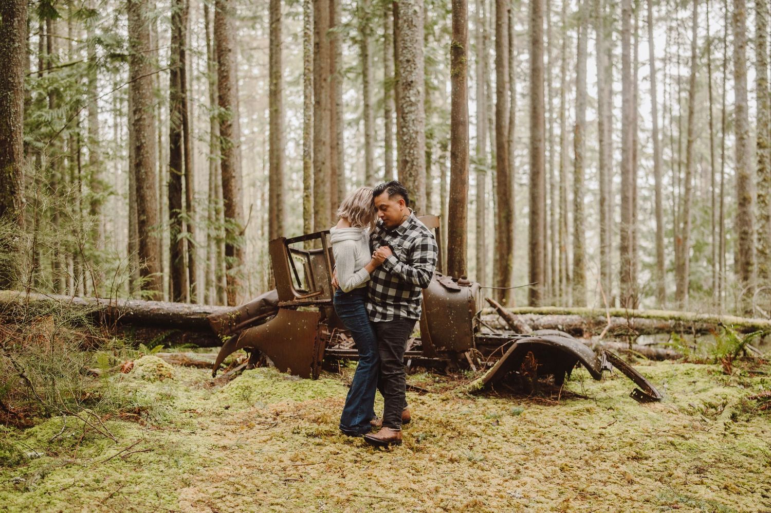 A rusty abandoned vintage car sits among tall evergreen trees in a mossy forest setting.