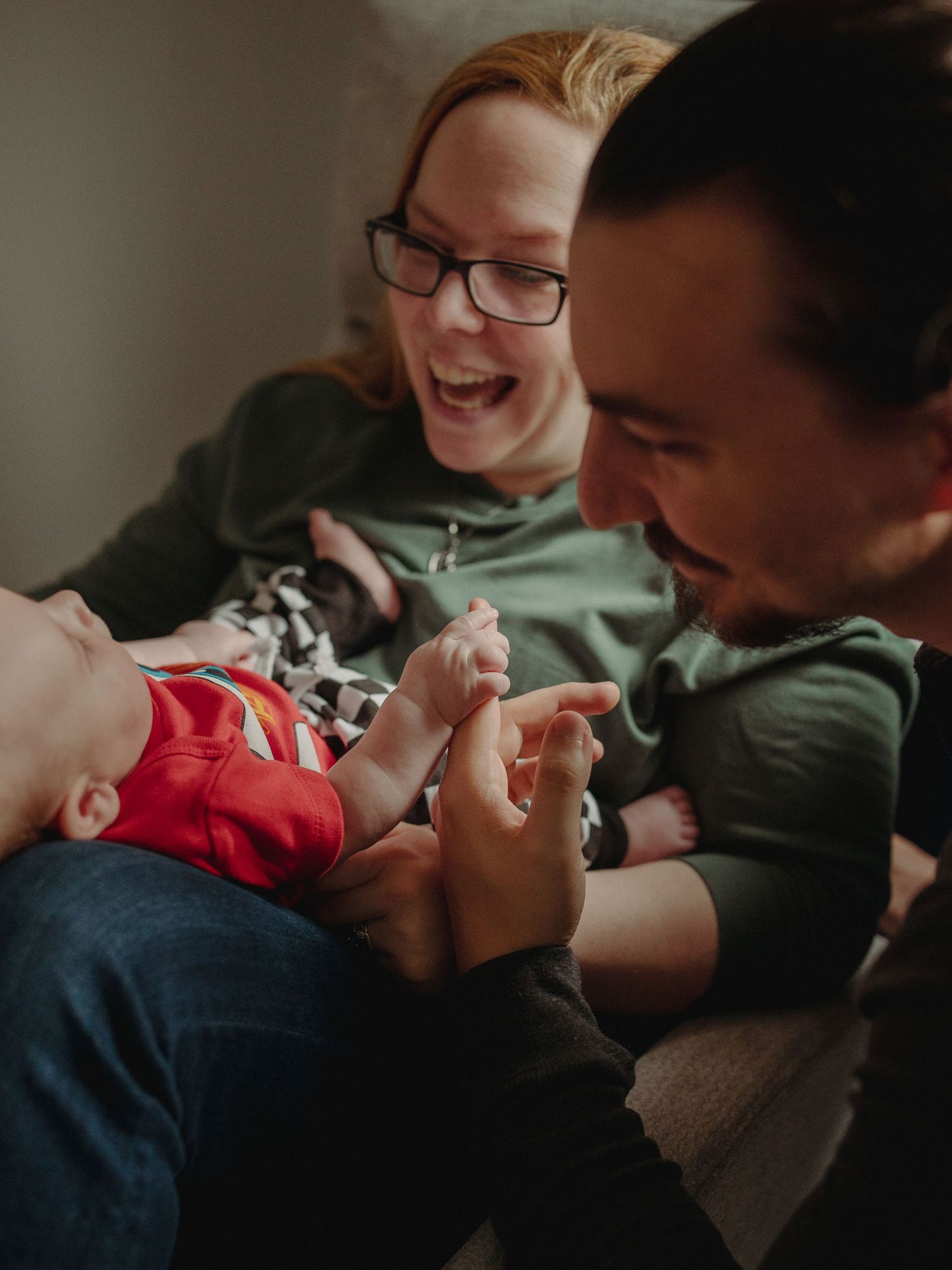 Happy parents laughing and playing with their newborn baby in red outfit during a cozy family moment at home.