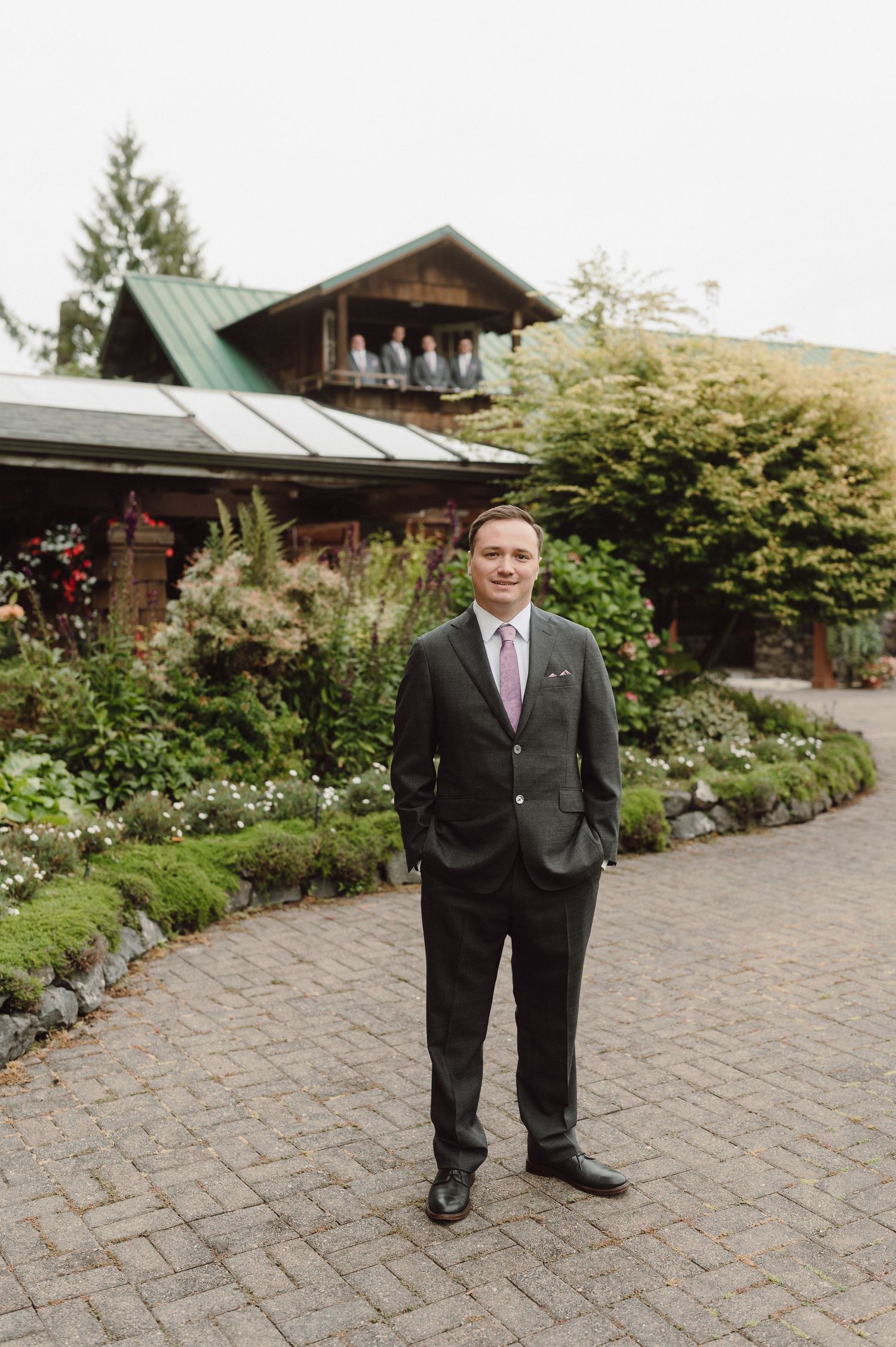 A person in a black suit stands on a brick patio outside a rustic wooden lodge surrounded by landscaped gardens.