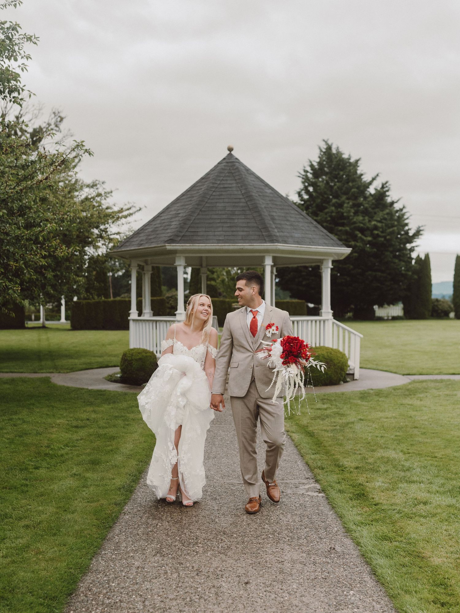 A couple walks down a path after their wedding ceremony in front of a white gazebo on a cloudy day.