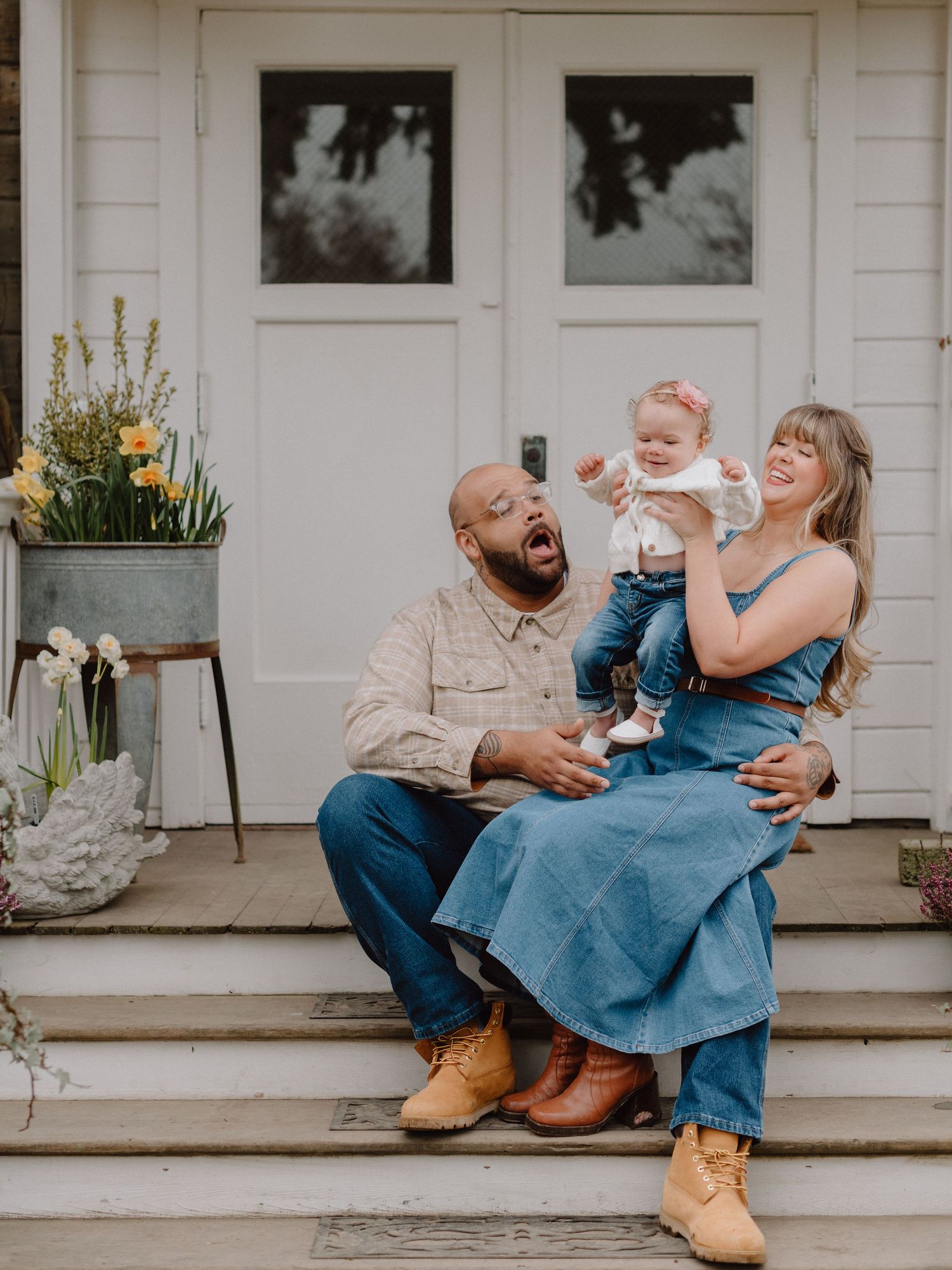 A joyful interracial family sits on white farmhouse porch steps, parents laughing with baby girl in denim outfits and tan boots.