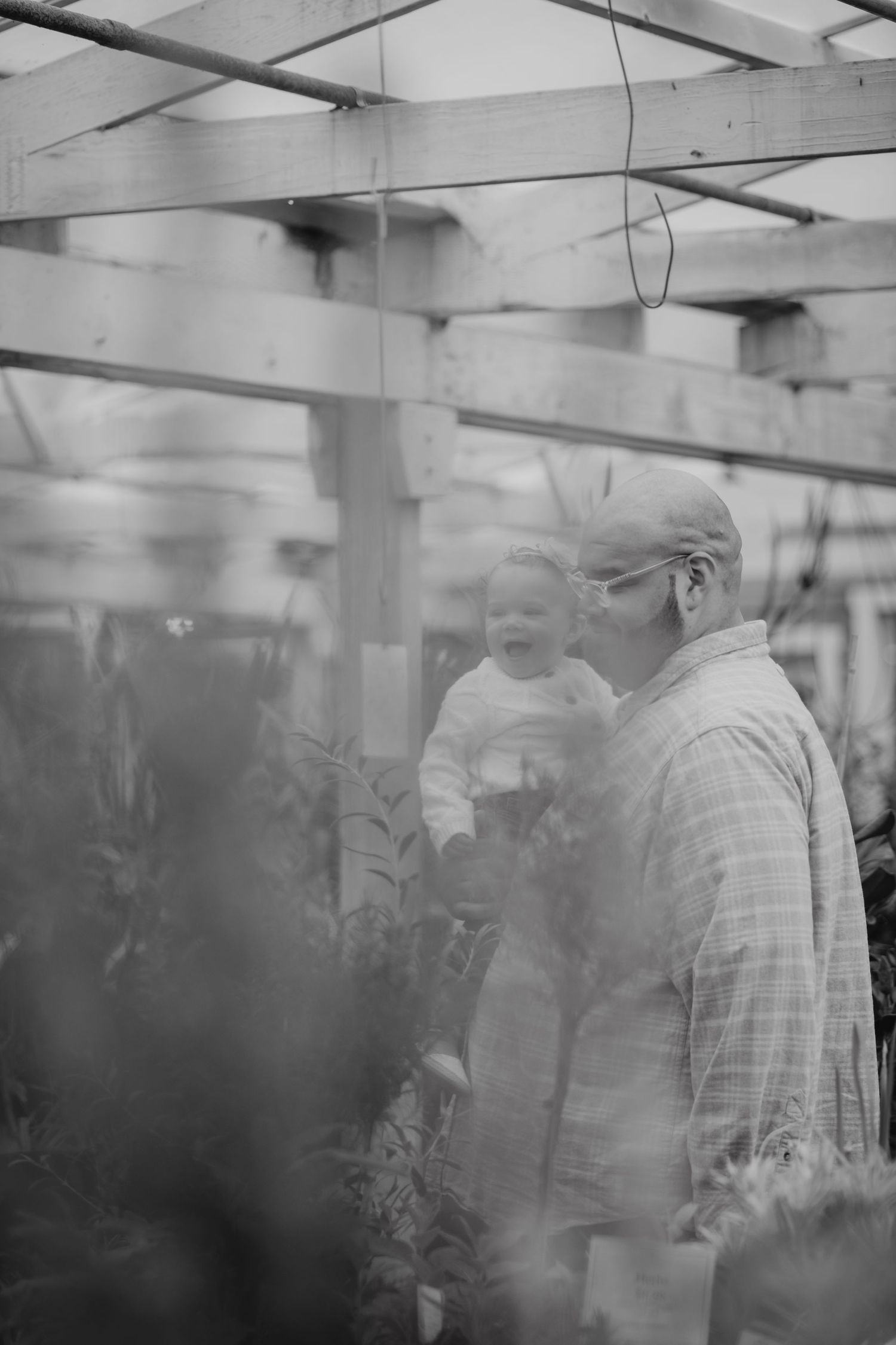 Black and white photo of an elderly man holding a smiling baby in a greenhouse surrounded by plants.