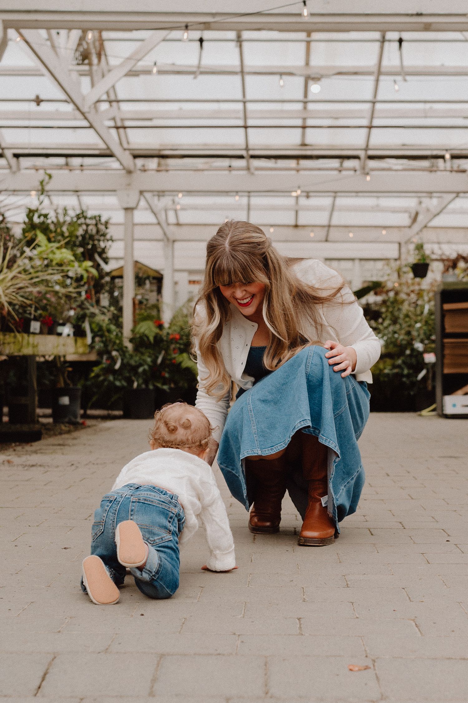 A smiling woman crouches beside a crawling baby in a sunlit greenhouse, both wearing denim outfits.